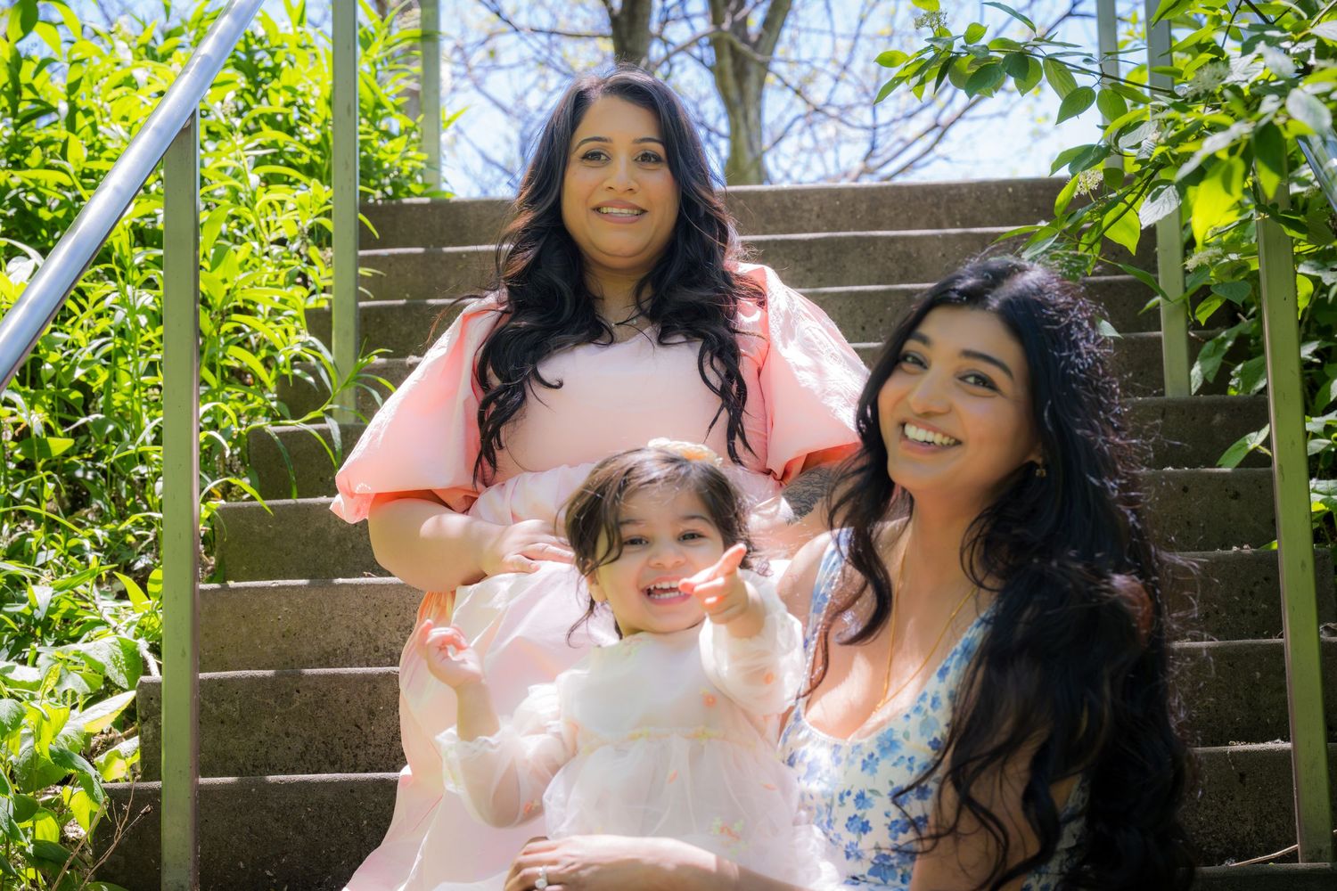 Woman in pink dress sits on steps with two younger girls in sunny outdoor setting.