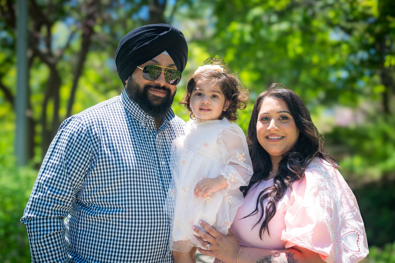 Sikh family portrait with father in blue turban, mother in pink dress, holding toddler in white outdoors.