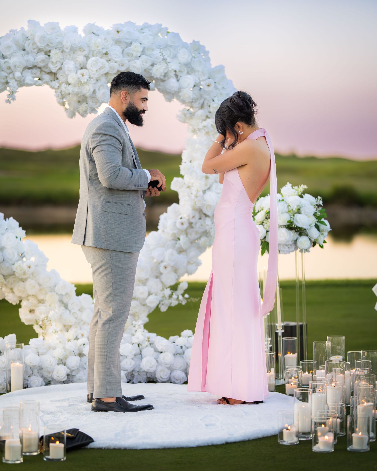 Romantic proposal setup with white floral arch and candles at sunset by a lake.