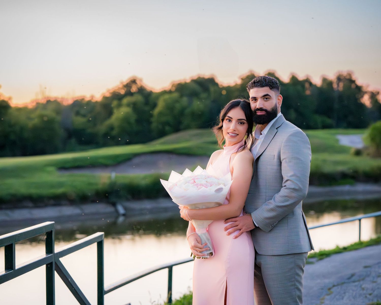 A couple in pink dress and gray suit embrace on a wooden boardwalk at sunset near a lake.