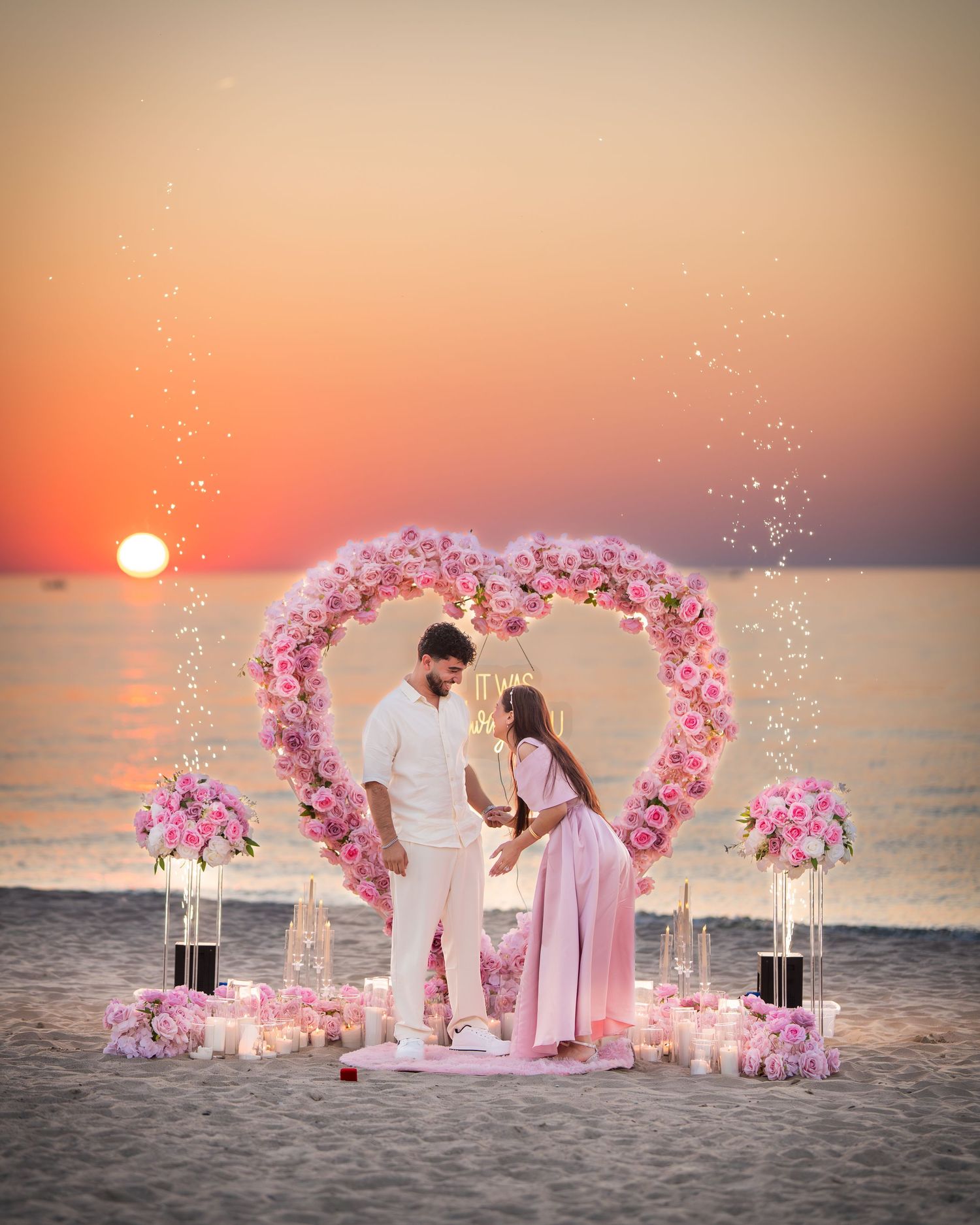 Beachside proposal scene with pink floral arch and glowing sunset over ocean.