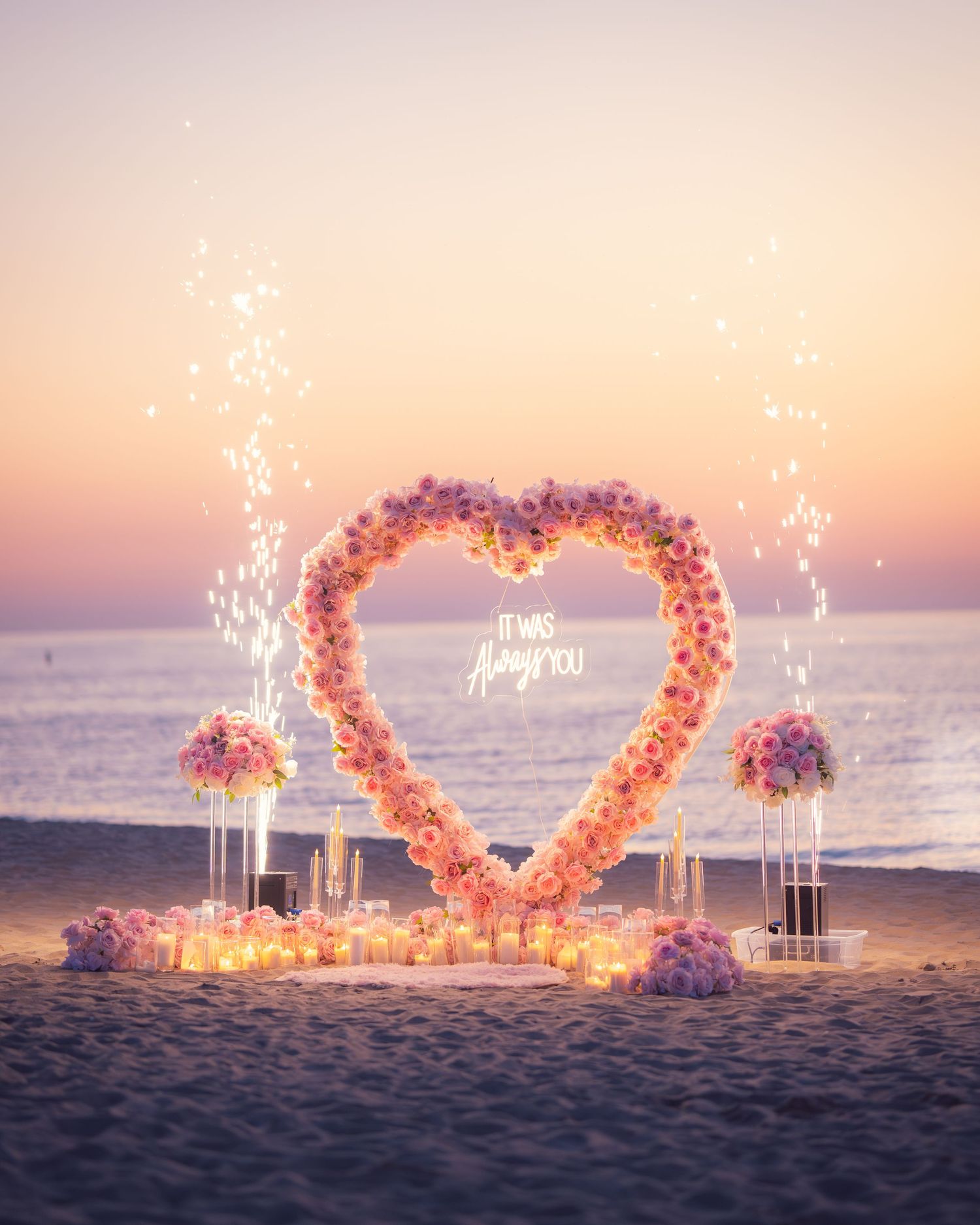 Magical beach proposal display with heart-shaped flower arch and sparkling lights at dusk.
