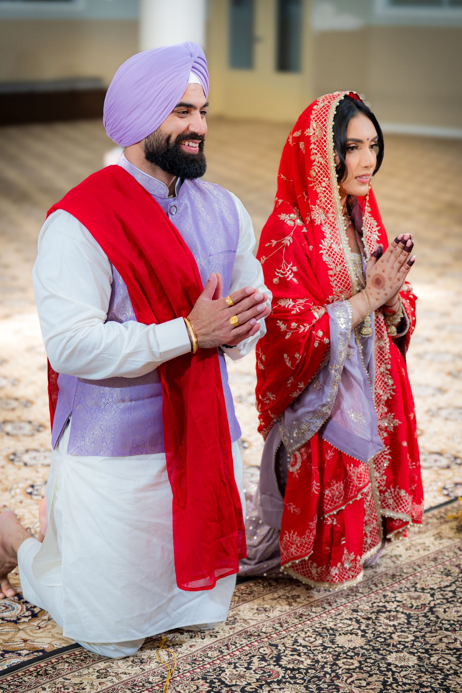 Sikh bride and groom in traditional attire kneel in prayer during wedding ceremony, wearing red and purple outfits.