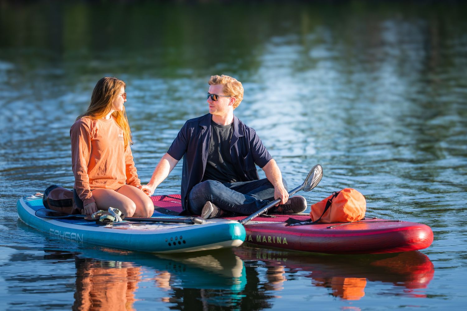 Young couple sitting on paddleboards on a reflective lake, sharing a quiet conversation at sunset.