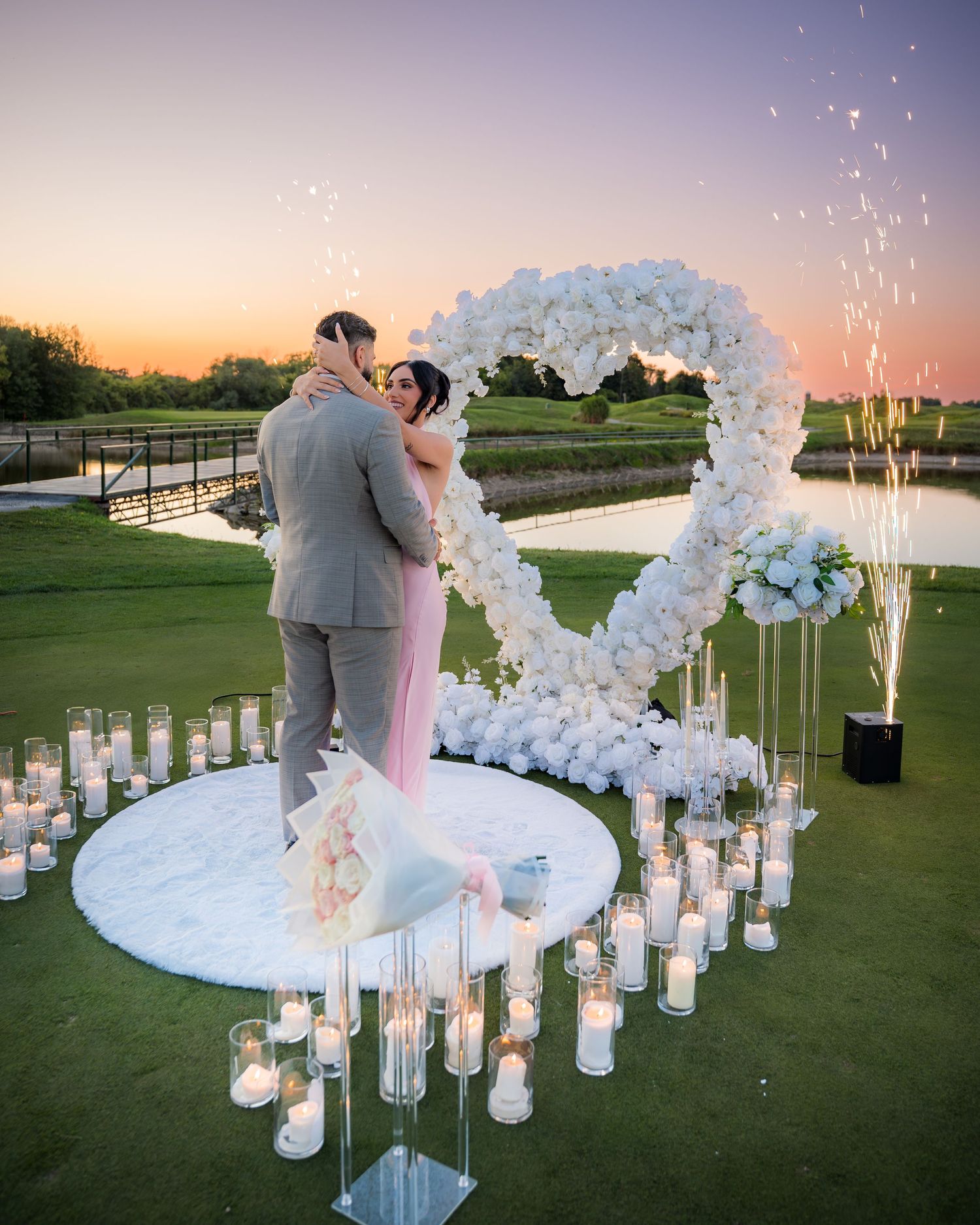 Romantic proposal setup with white floral arch and glowing candles on a golf course.