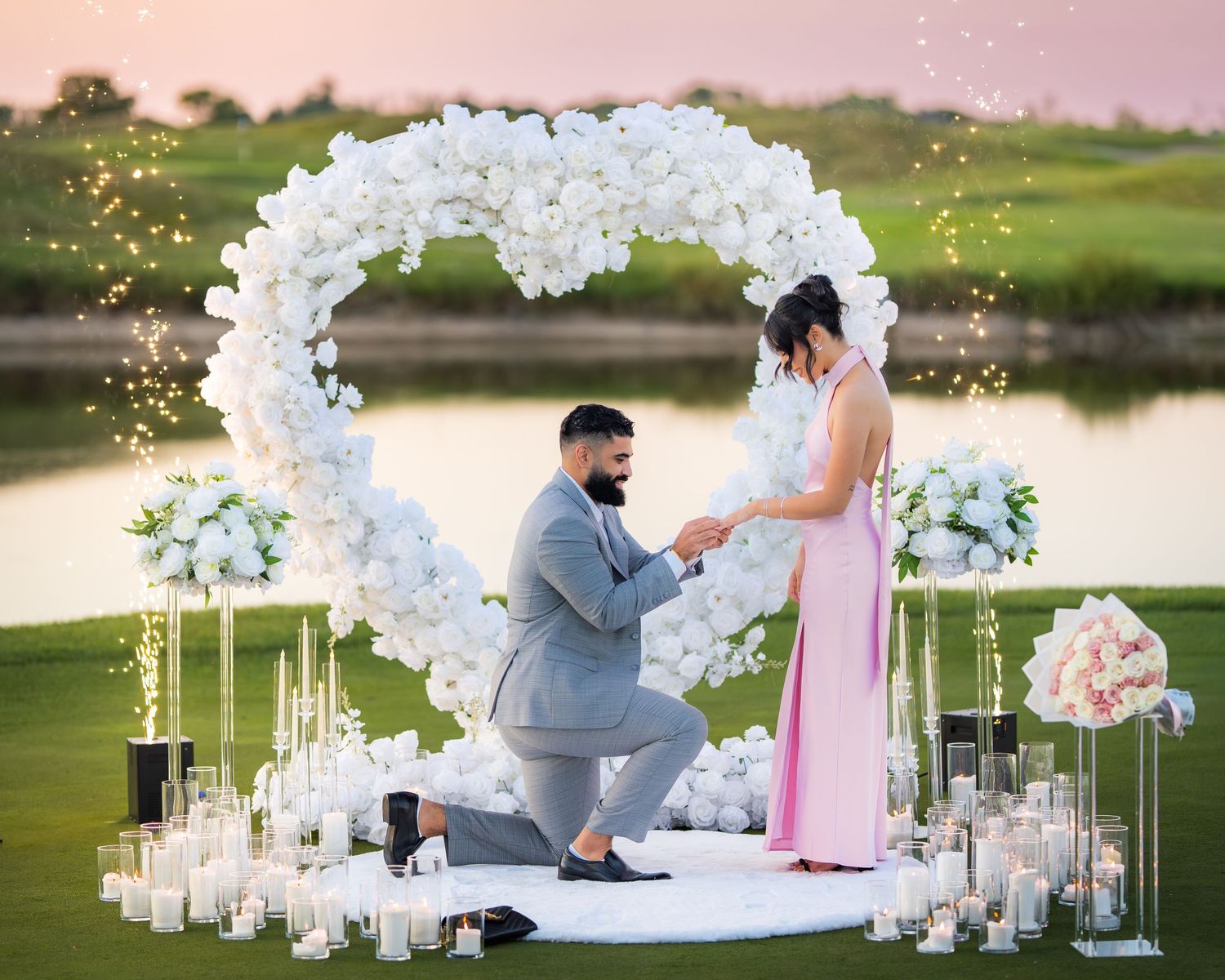 Dreamy outdoor proposal setup with white floral arch and glowing candles at dusk.