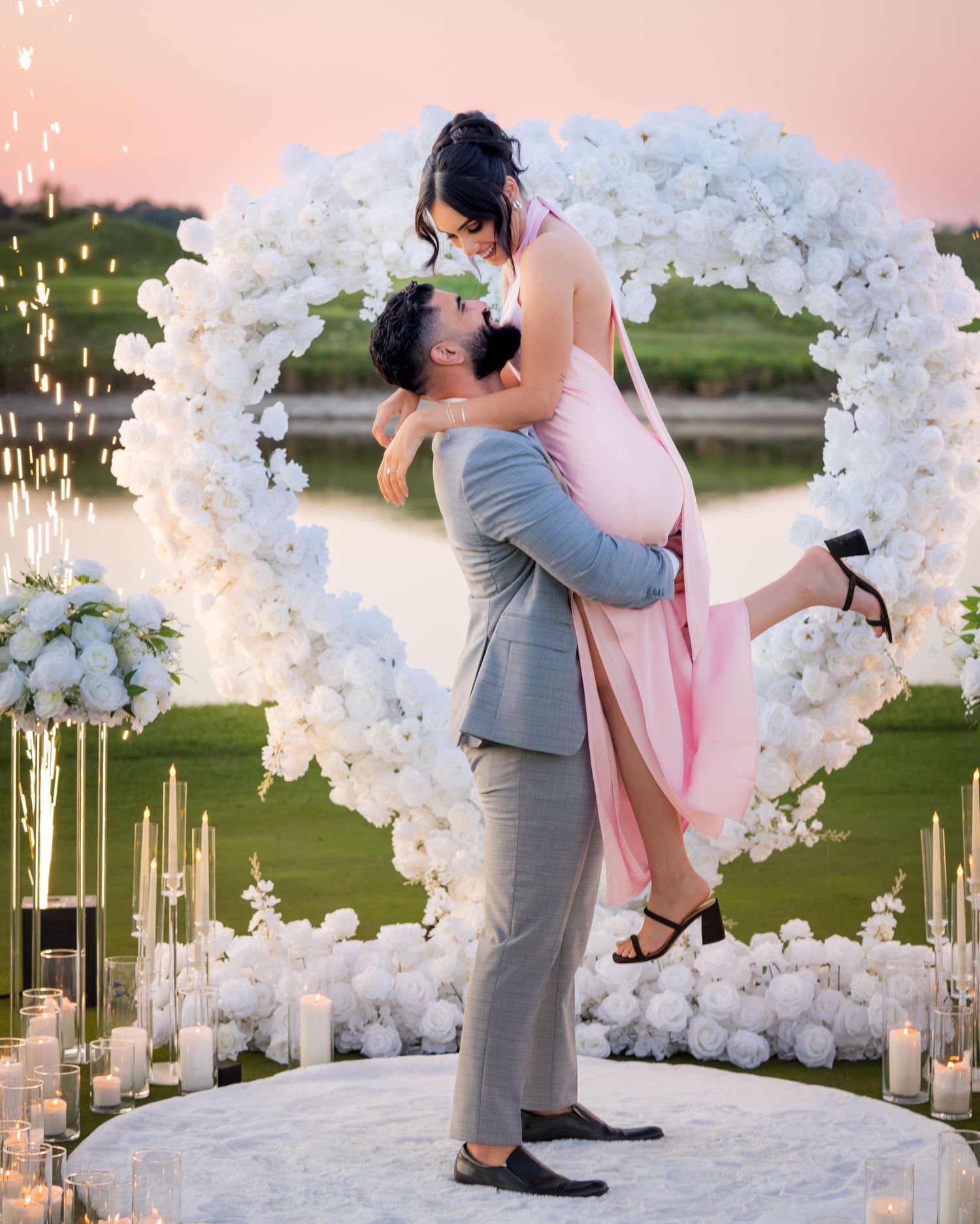 Couple embracing beneath an elegant white balloon and floral arch with glowing candles at dusk.