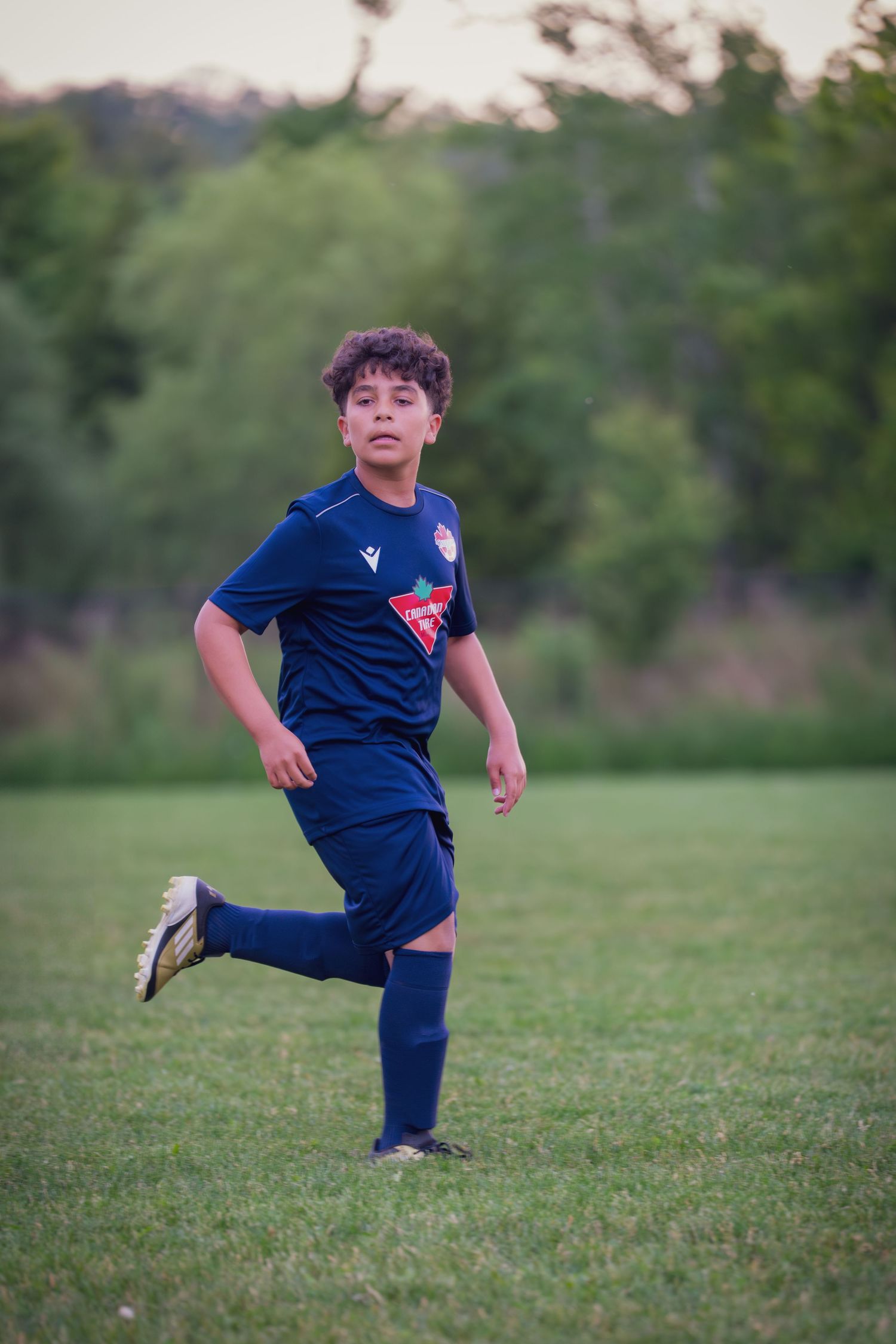 Young soccer player in navy blue uniform practices footwork drills on a grassy field at sunset.