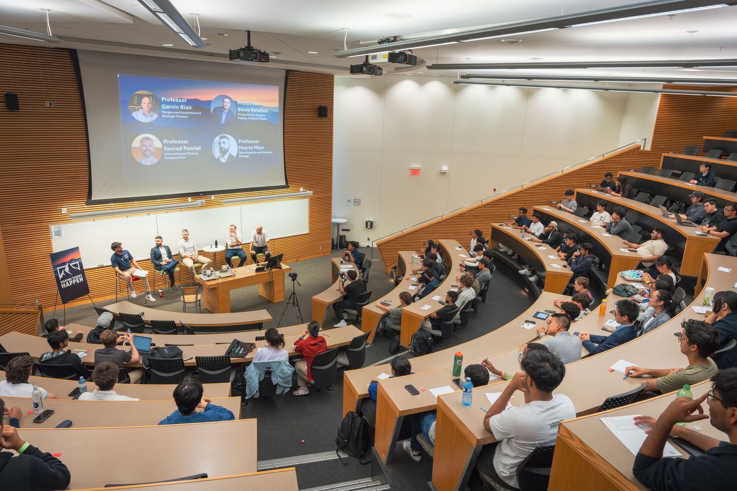 Students sit in a modern lecture hall with tiered seating while viewing presentations on multiple projector screens.