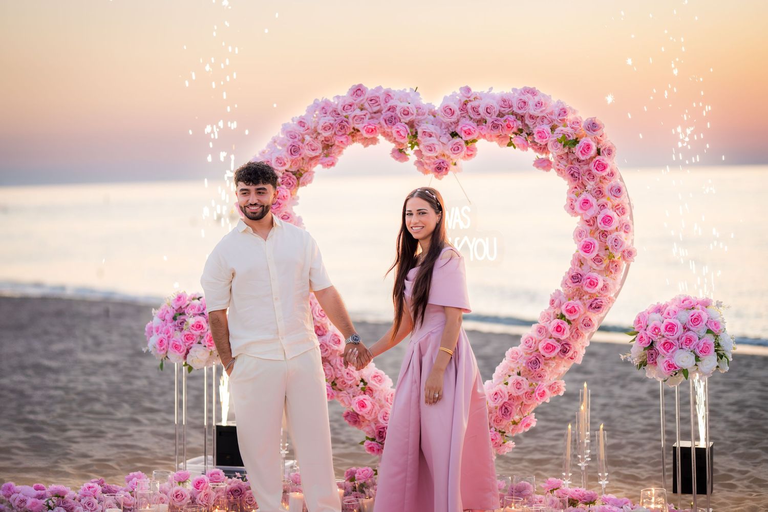 Romantic beach proposal setup with pink floral heart arch and candles at sunset.