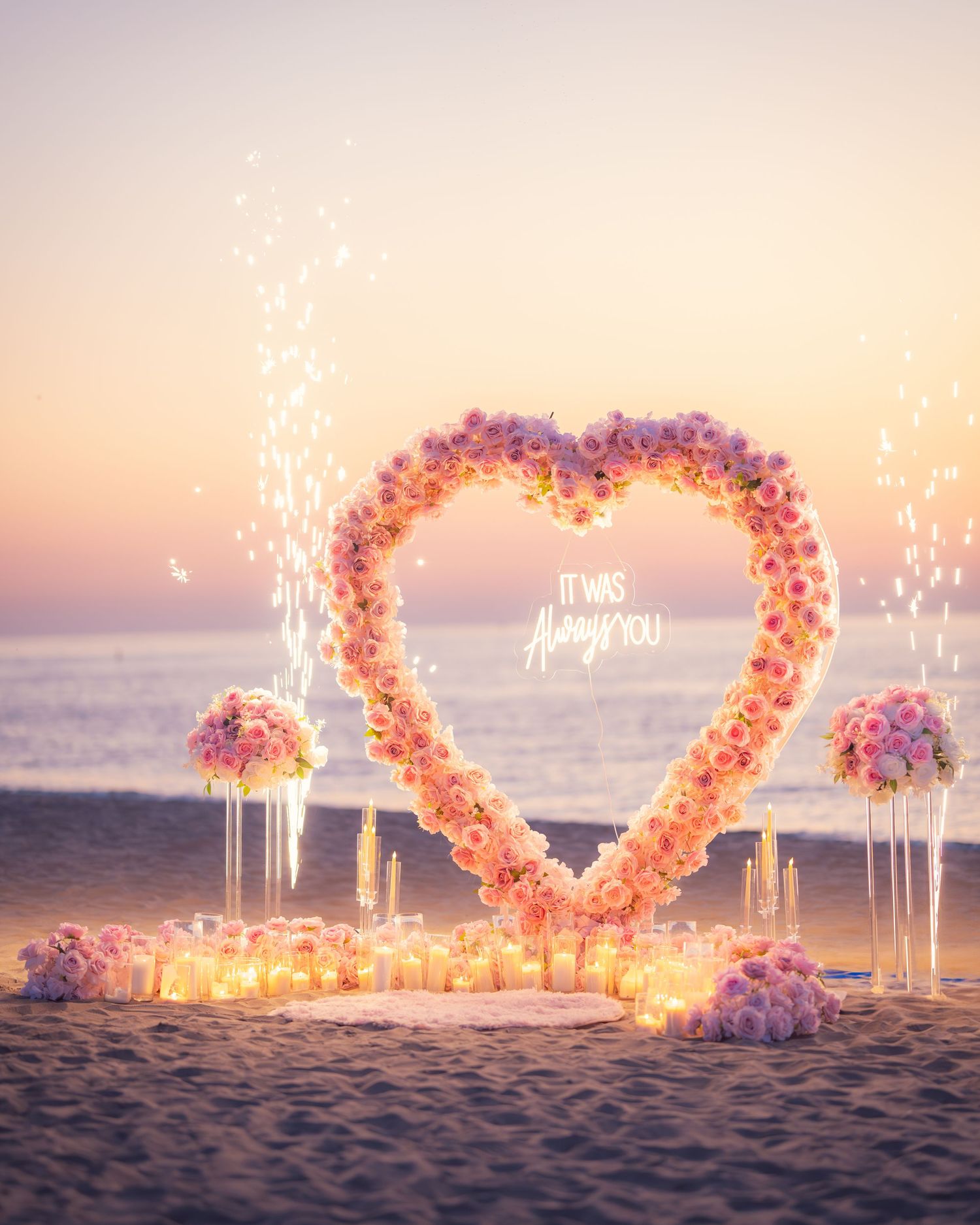 Romantic beach setup with pink floral heart arch and illuminated candles at golden hour.