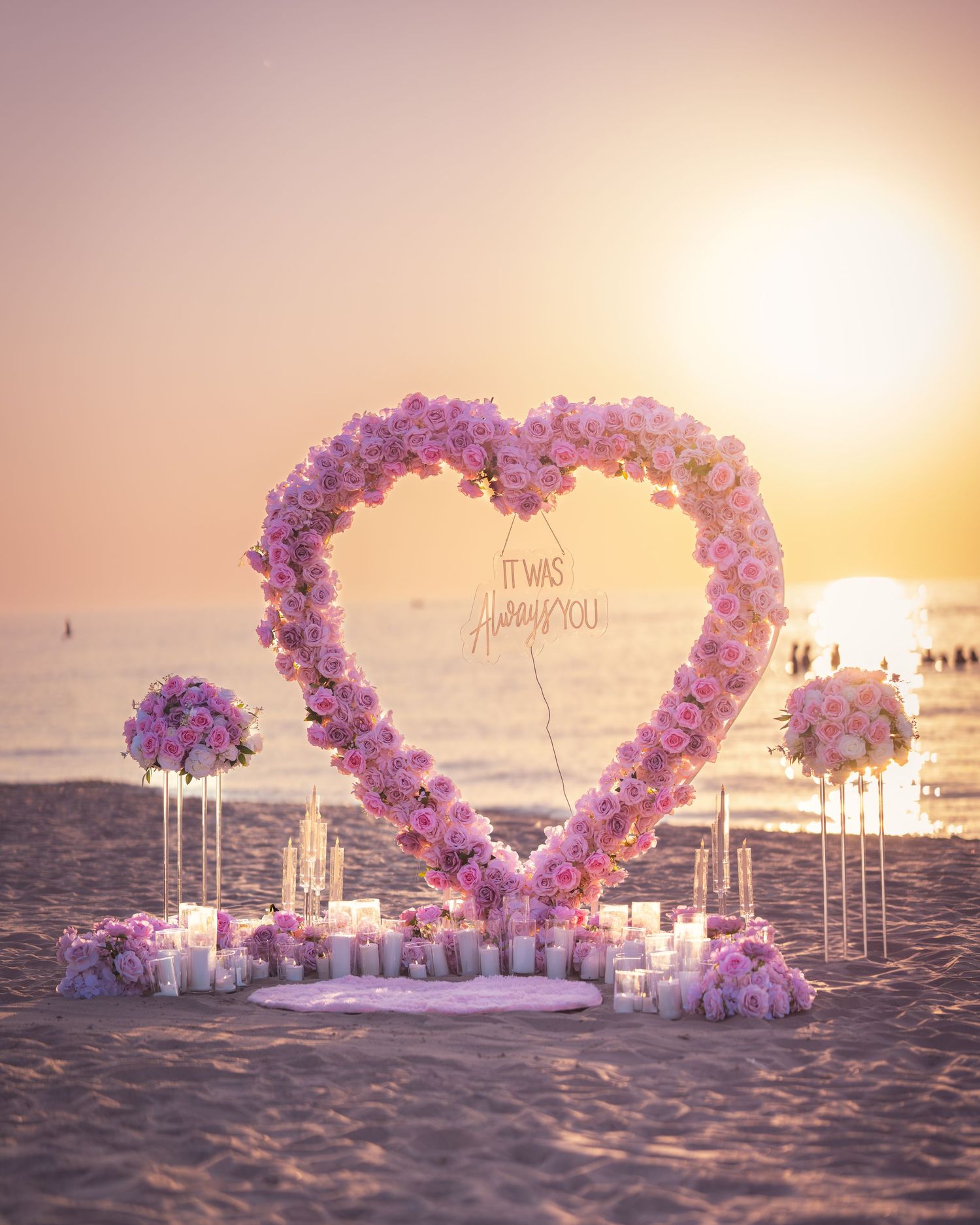 Pink and purple balloon arch in heart shape decorates a beach during golden hour.