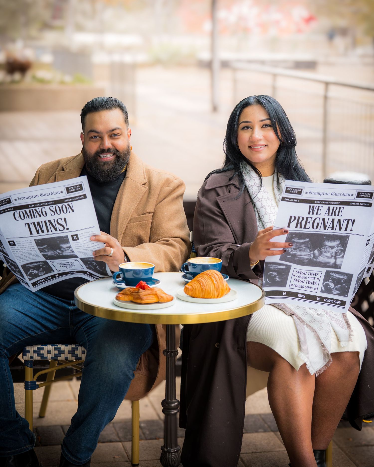 A couple sits at an outdoor cafe table holding custom newspapers announcing their pregnancy.