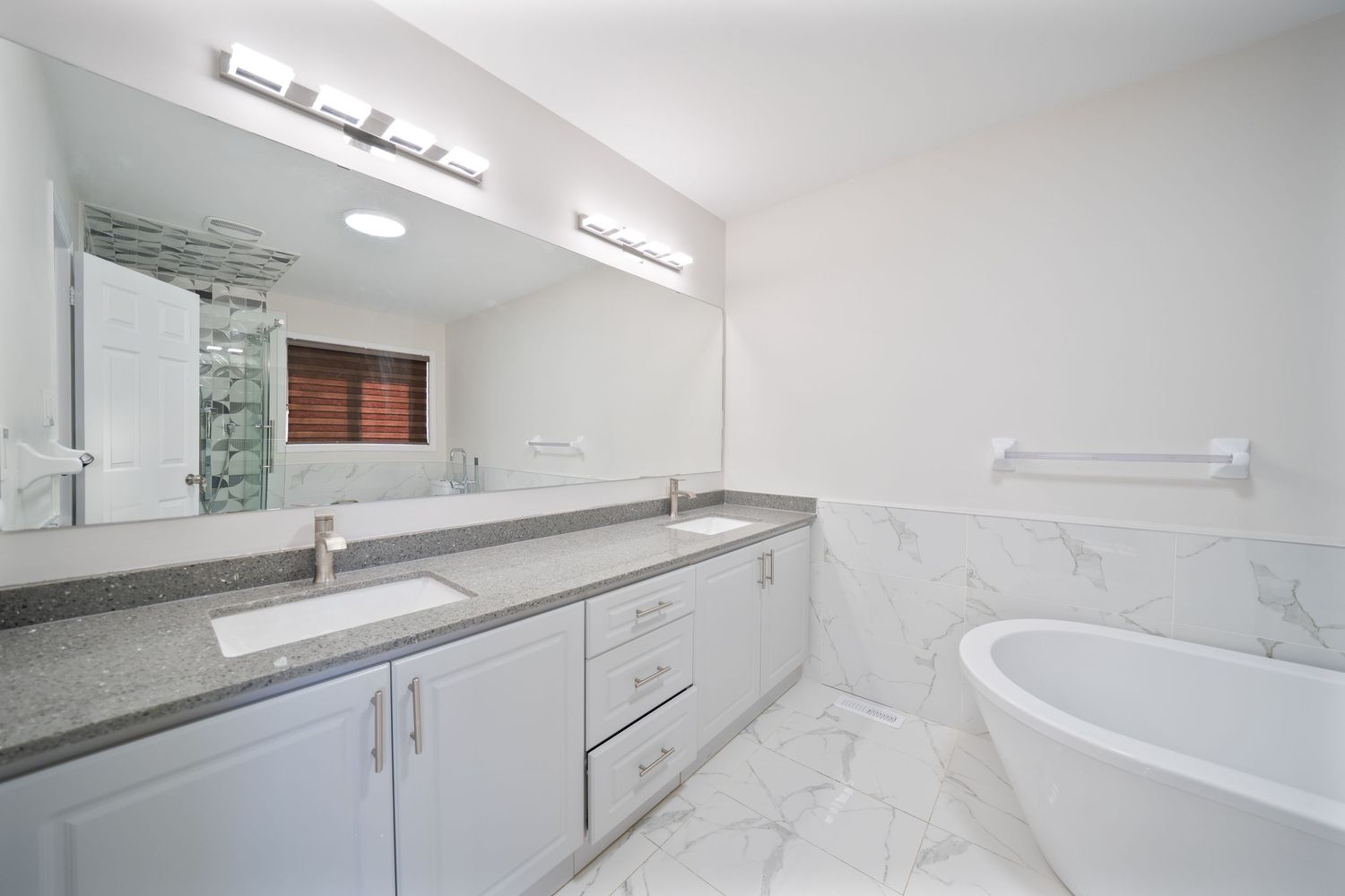 Modern white bathroom interior with marble floors, freestanding tub, and long double vanity with granite countertop.