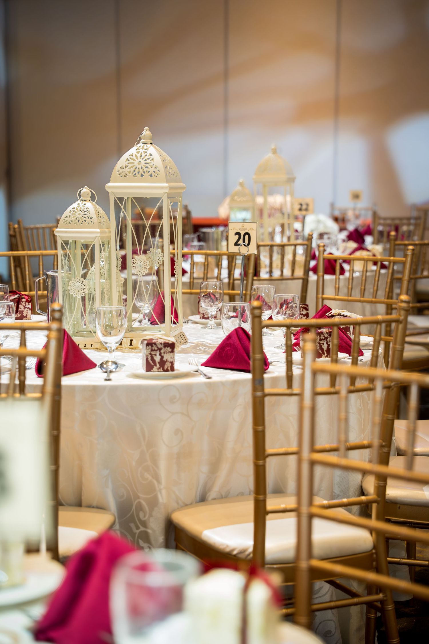 Elegant wedding reception decor with gold lantern centerpieces and burgundy accents on white tablecloths.