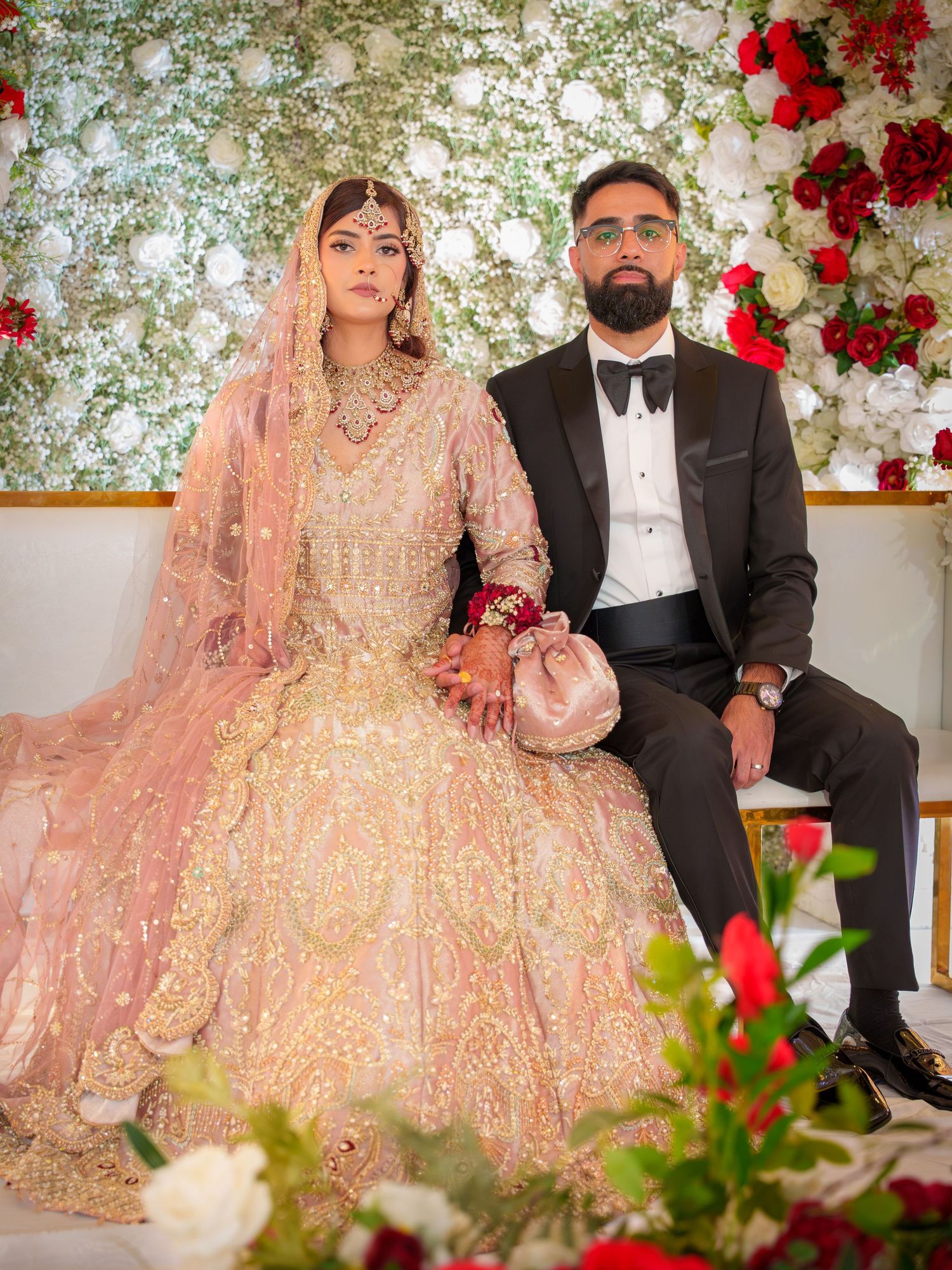South Asian wedding couple poses against floral wall with red roses and white flowers.