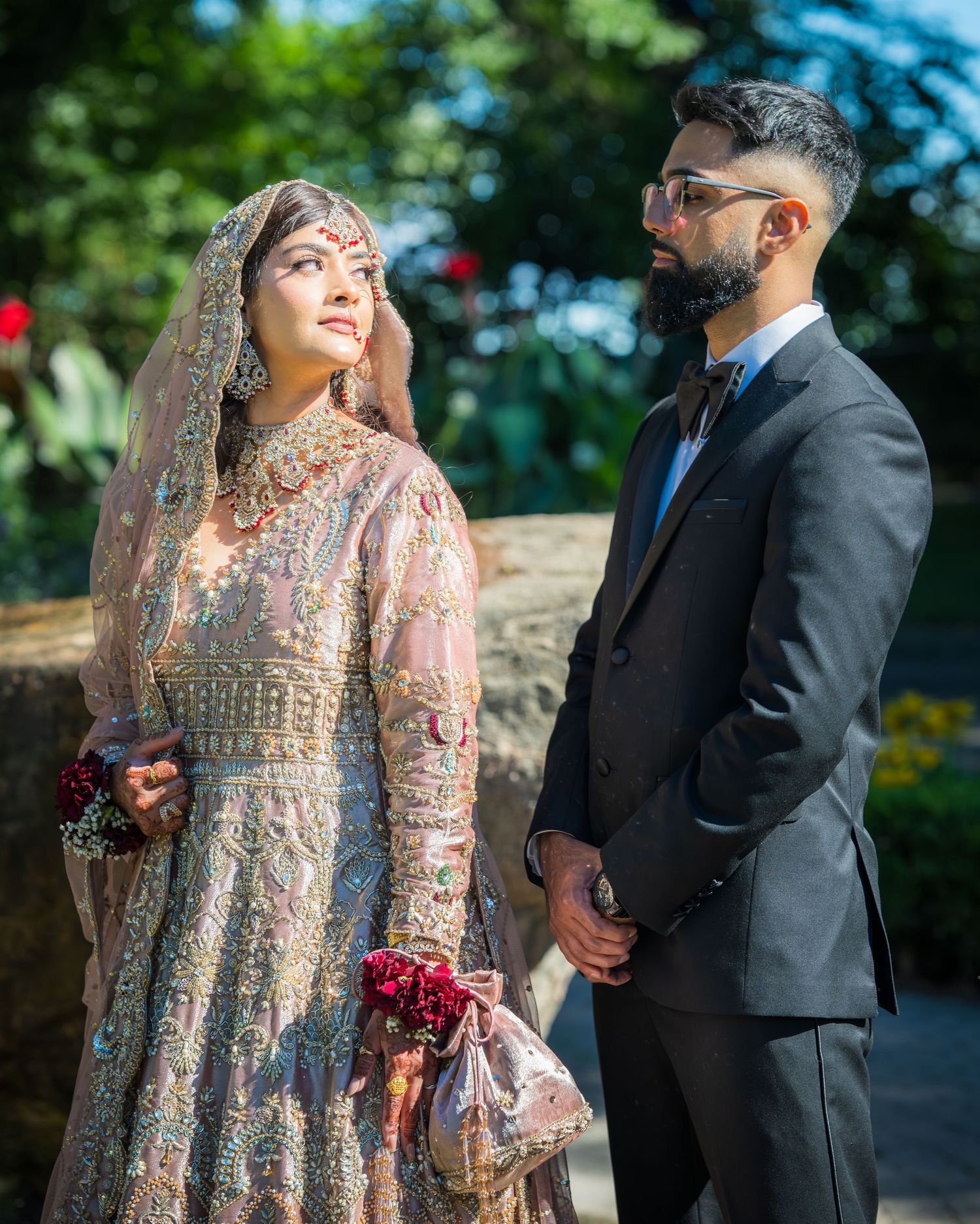 Couple in traditional South Asian wedding attire pose together in an outdoor garden setting during their special day.