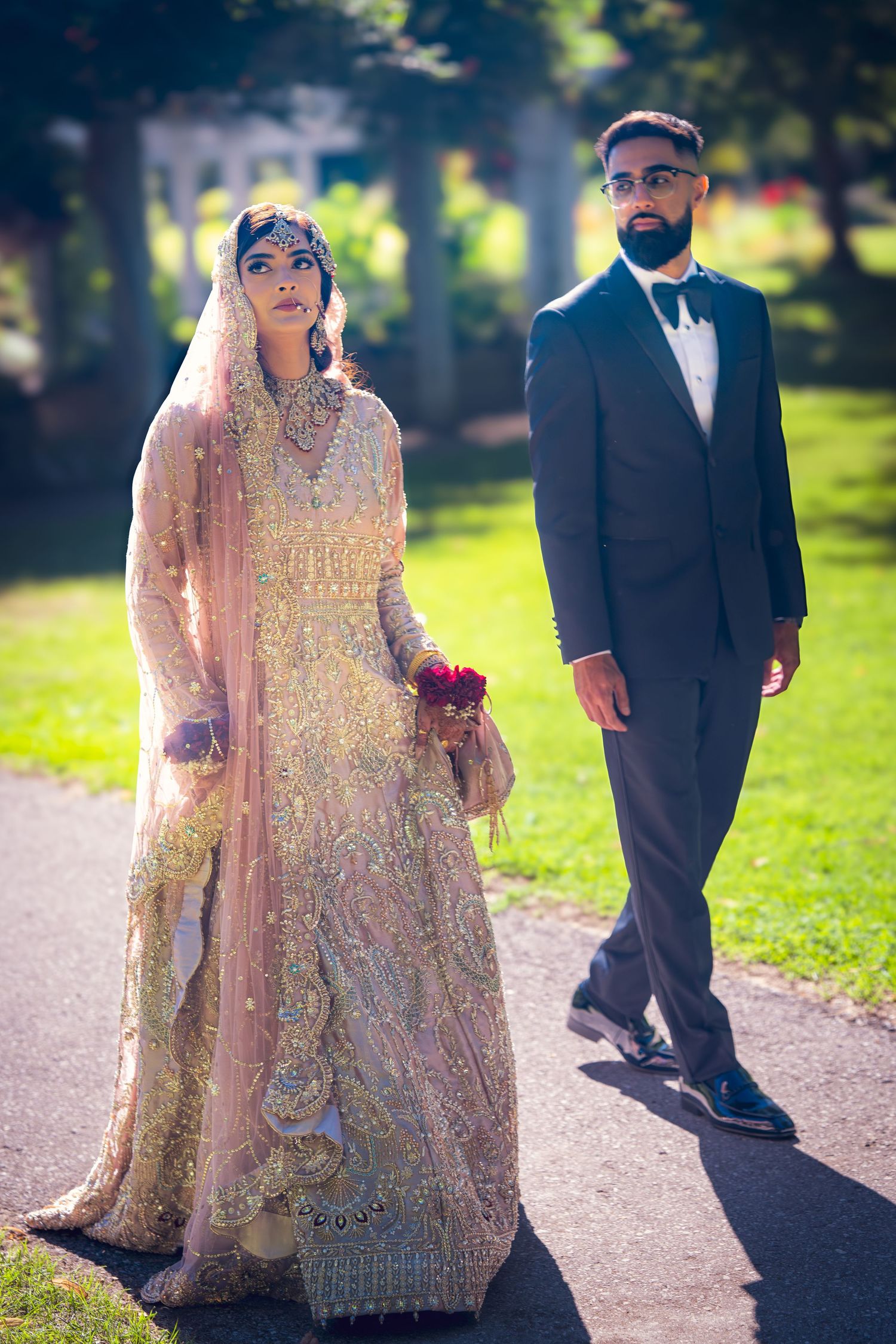 Wedding couple in traditional South Asian attire walking along garden path during photoshoot.