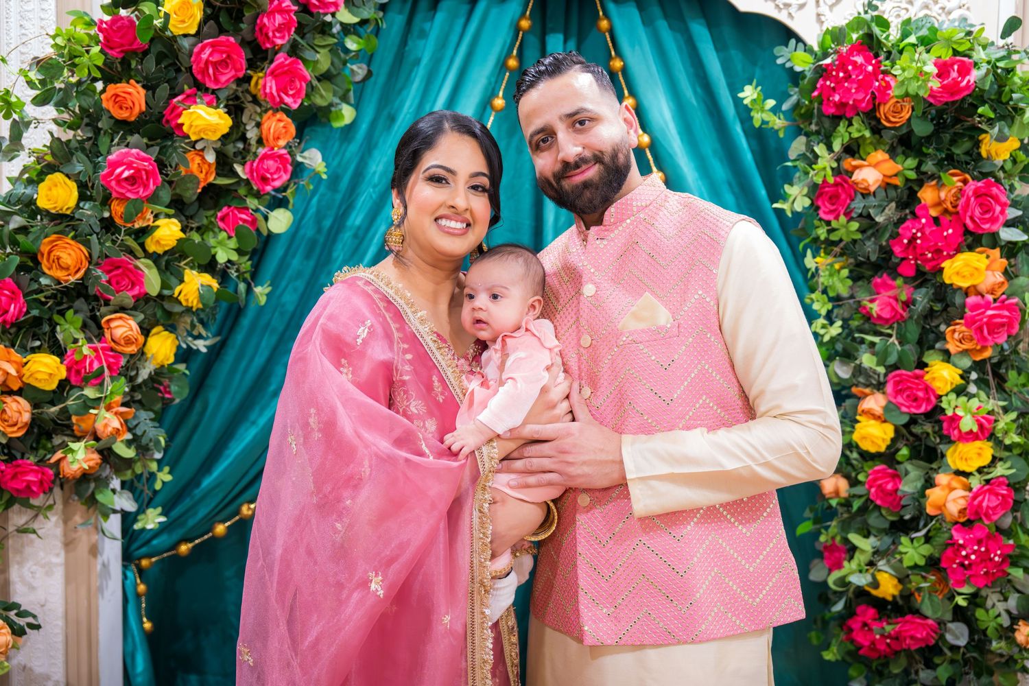 Indian family in pink traditional attire poses with baby in front of colorful floral arch at celebration.