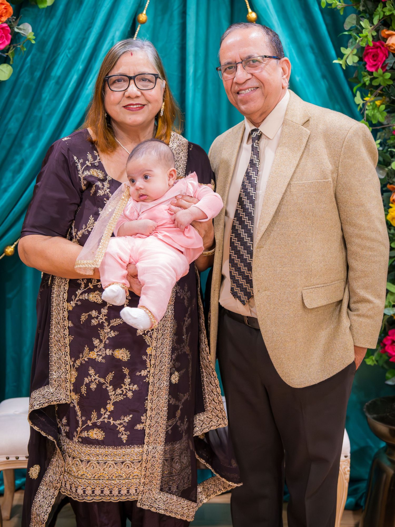 Grandparents in traditional South Asian attire holding a baby in pink at a decorated event.