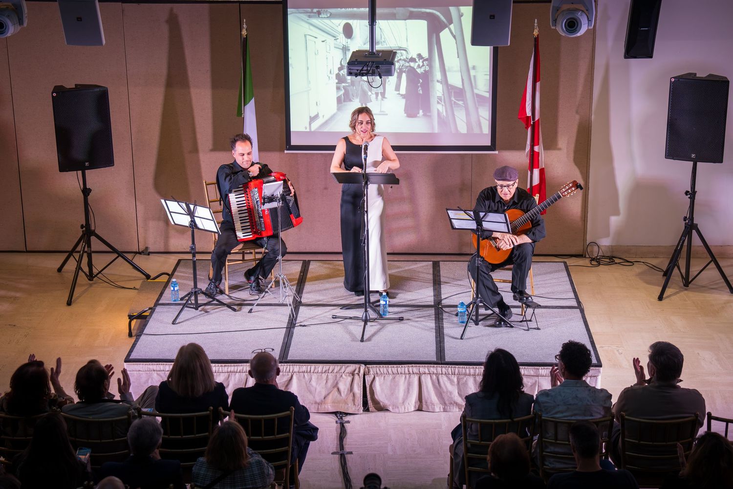A trio performs on stage with accordion, vocals, and guitar before a seated audience with flags displayed.