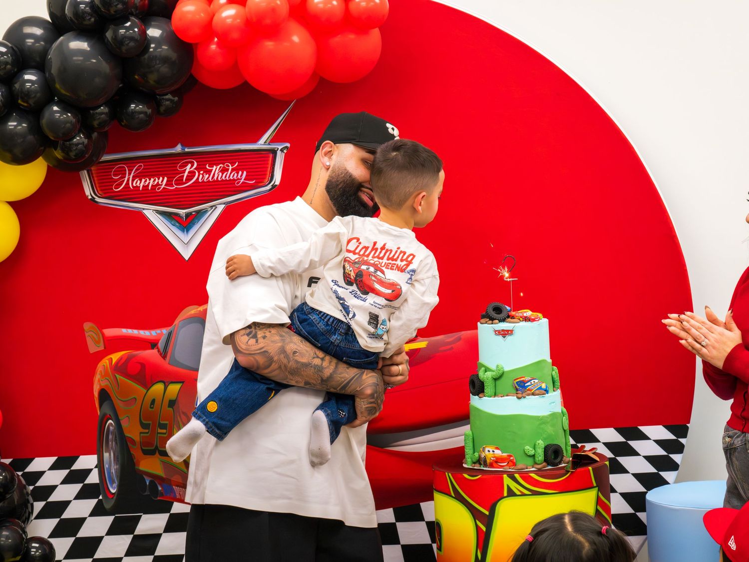 A tattooed man holds a young child near a Cars-themed birthday cake and balloon arch backdrop.