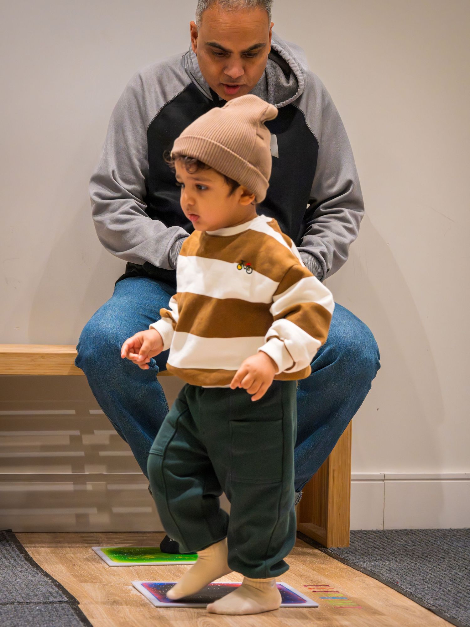 A man helps a toddler in a striped sweater and beanie hat stand on a colorful mat indoors.