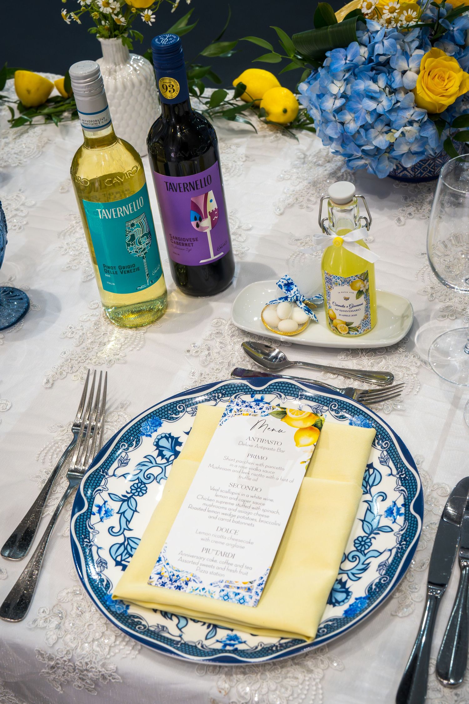 Elegant wedding table setting with blue and white plates, yellow napkins, menu card, and Tavernello wine bottles with floral centerpiece.