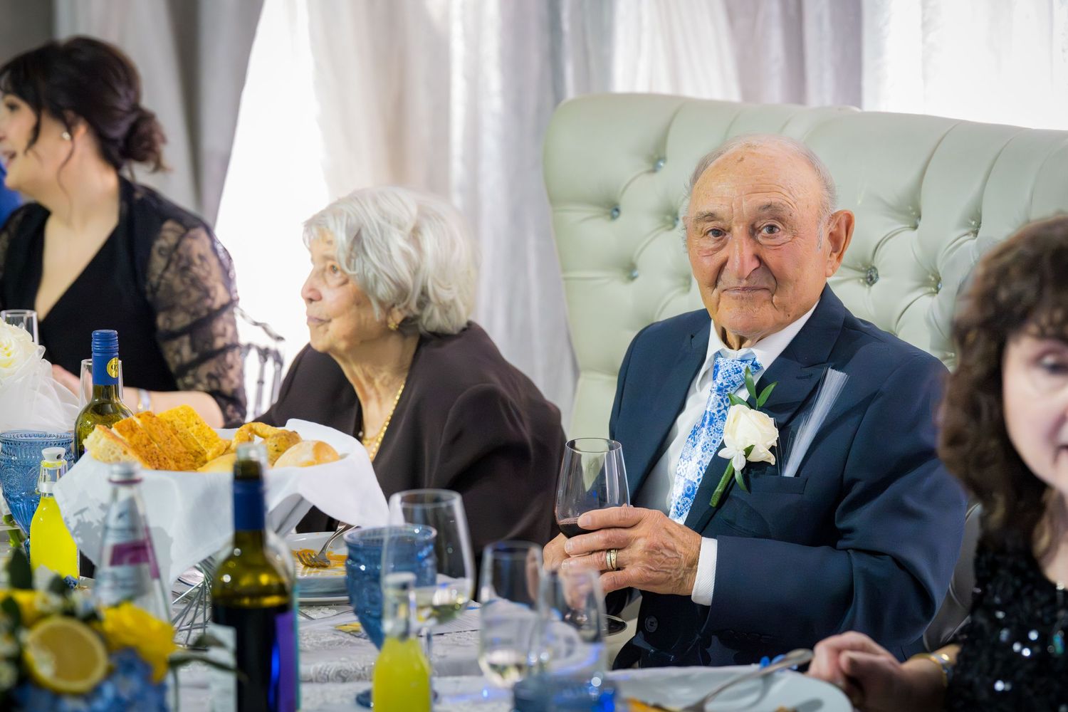 Elderly man smiling and holding red wine glass at festive anniversary dinner table with guests in background.