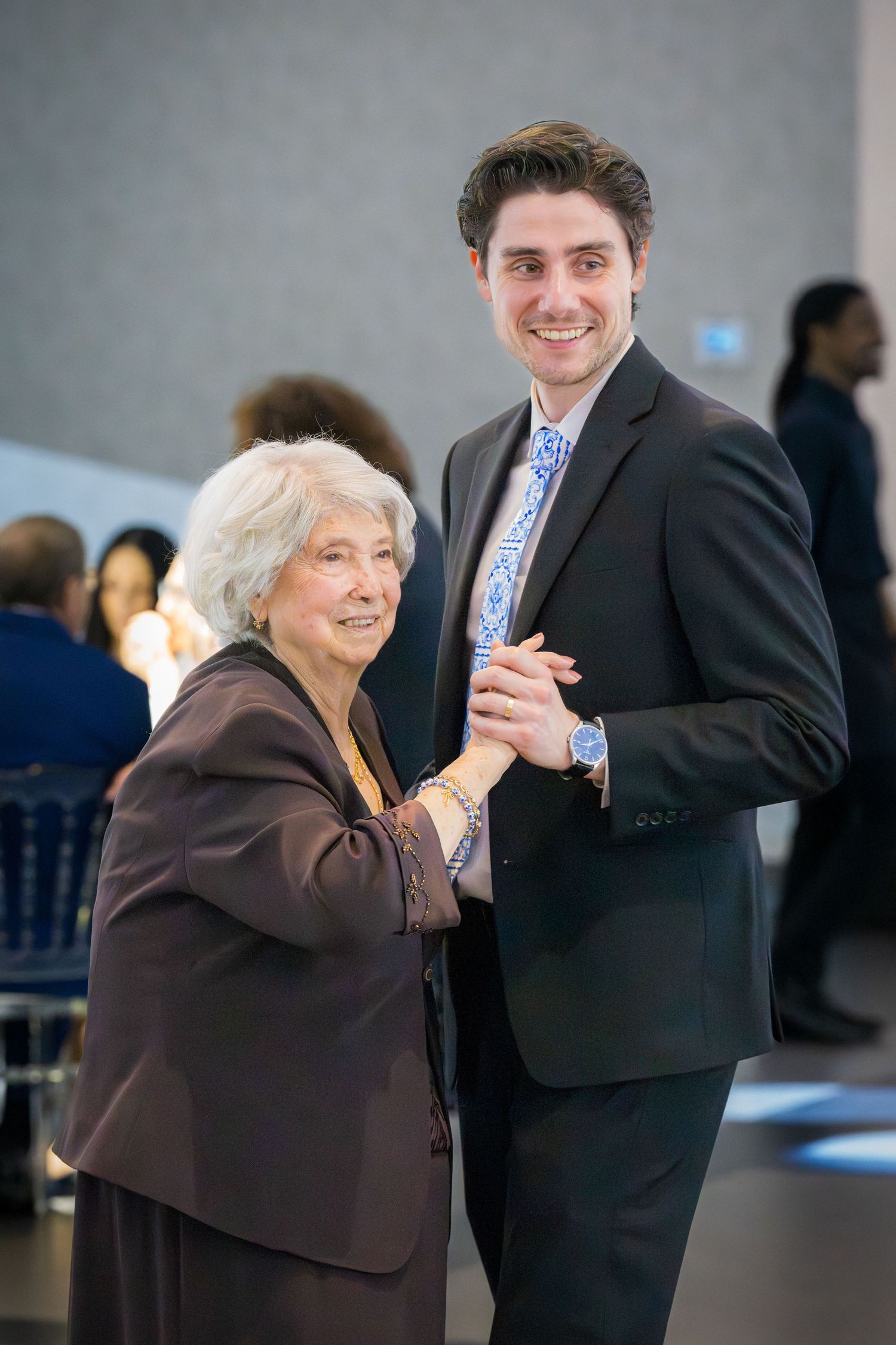 Elderly woman and young man in black suit dancing together at a formal celebration.
