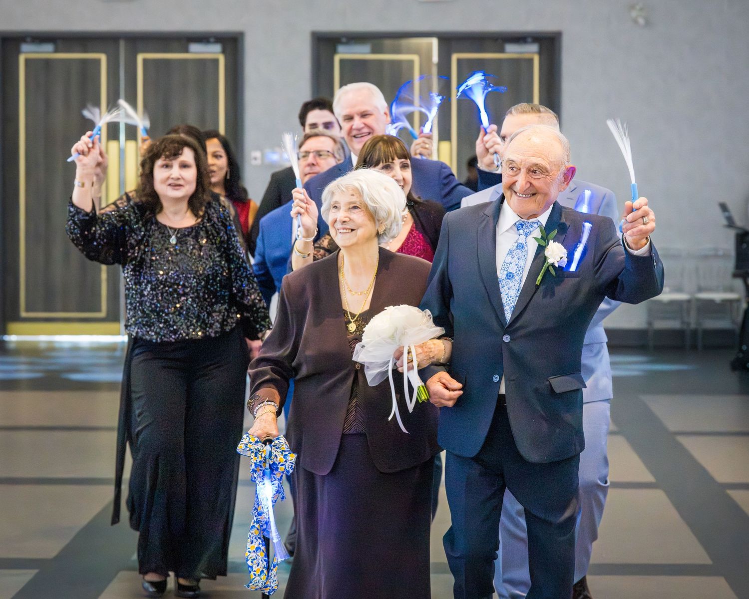 Elderly couple leads joyful procession holding sparklers at formal celebration event with smiling guests behind them.