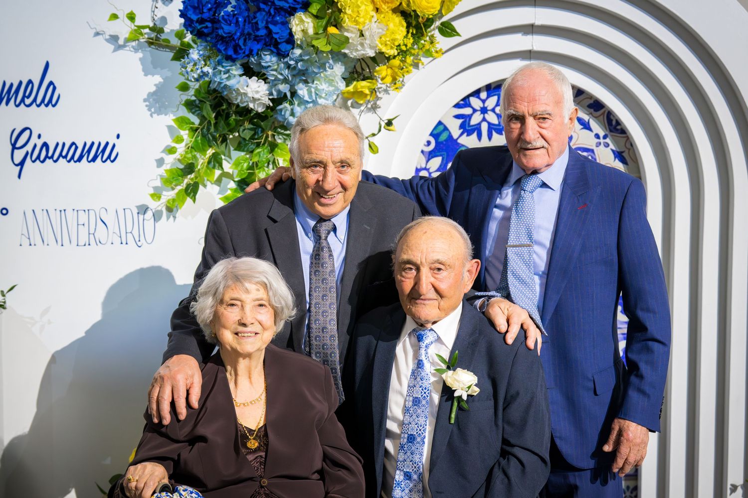 Elderly couple seated with two adult sons at Giovanni and Carmela anniversary celebration backdrop.