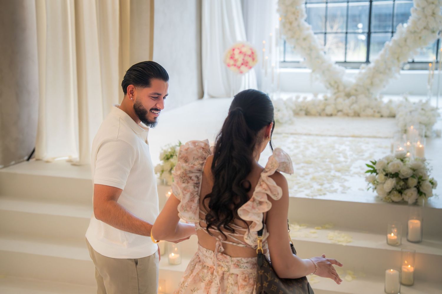 Man helping woman with floral dress near romantic white flower heart arch with candles.