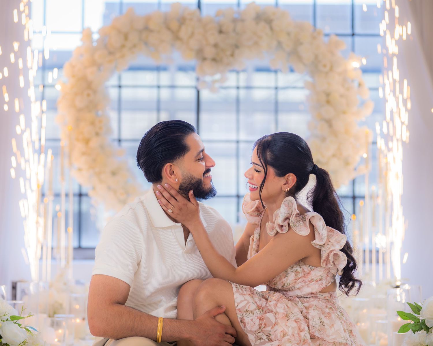 Couple sharing a tender moment under a glowing white floral heart arch during their engagement photoshoot.