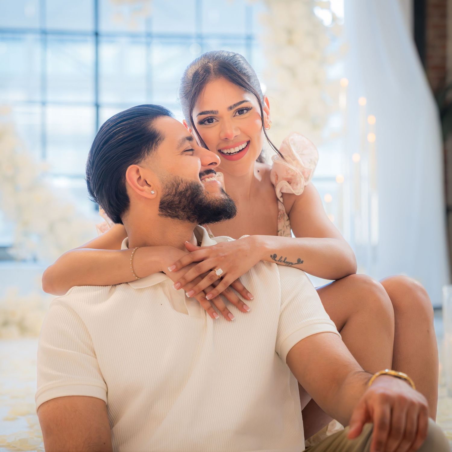 Happy couple sitting together smiling, woman hugging man from behind near white floral heart arch.