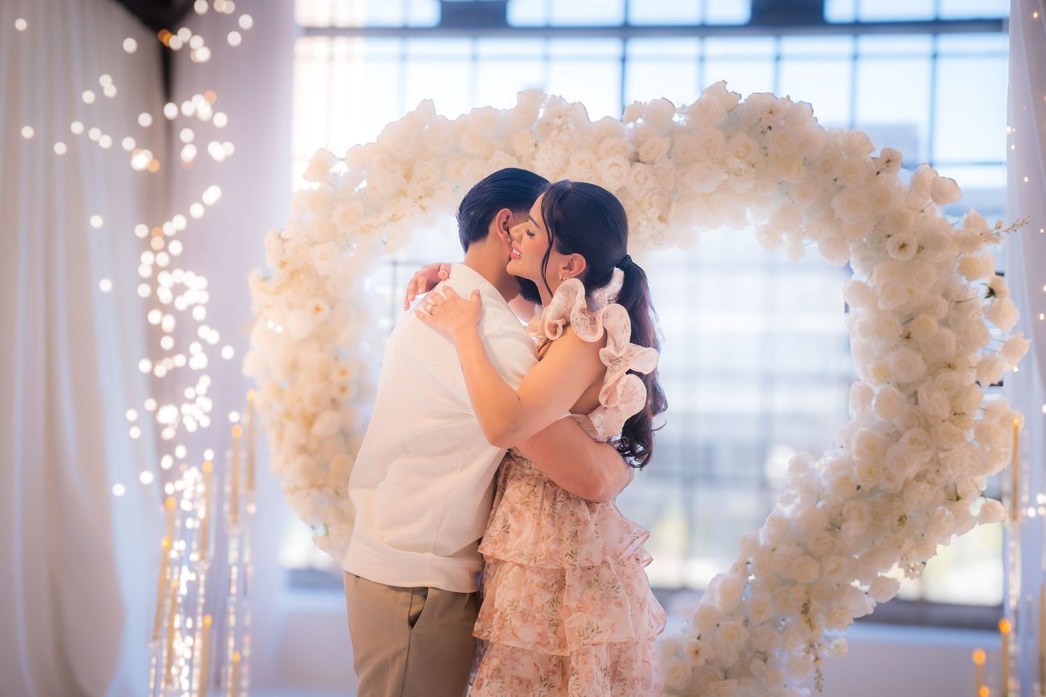 Couple sharing first kiss under white floral heart arch with twinkling fairy lights backdrop.