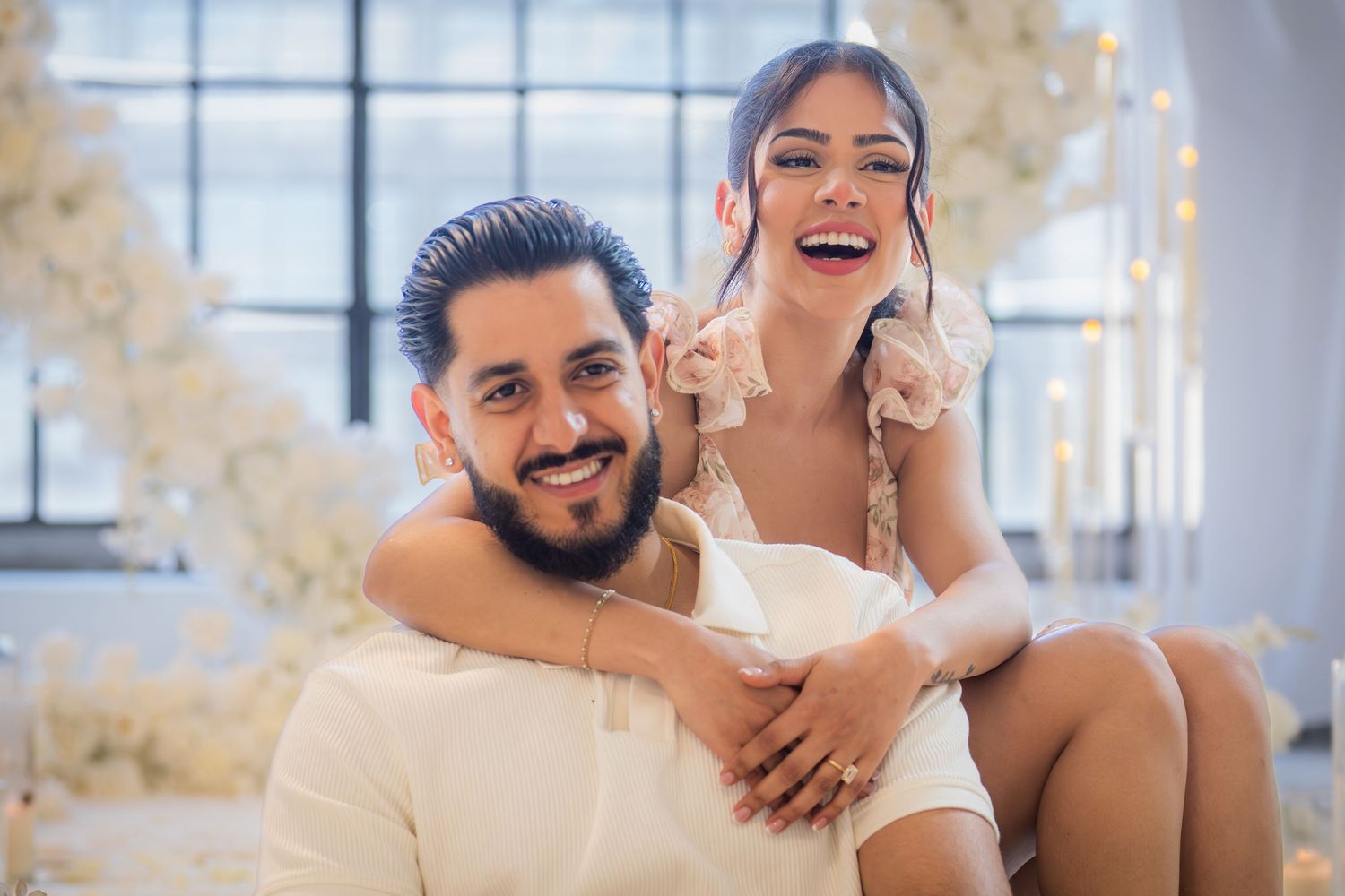 Joyful couple smiling at camera, woman sitting on mans lap surrounded by white floral decorations.