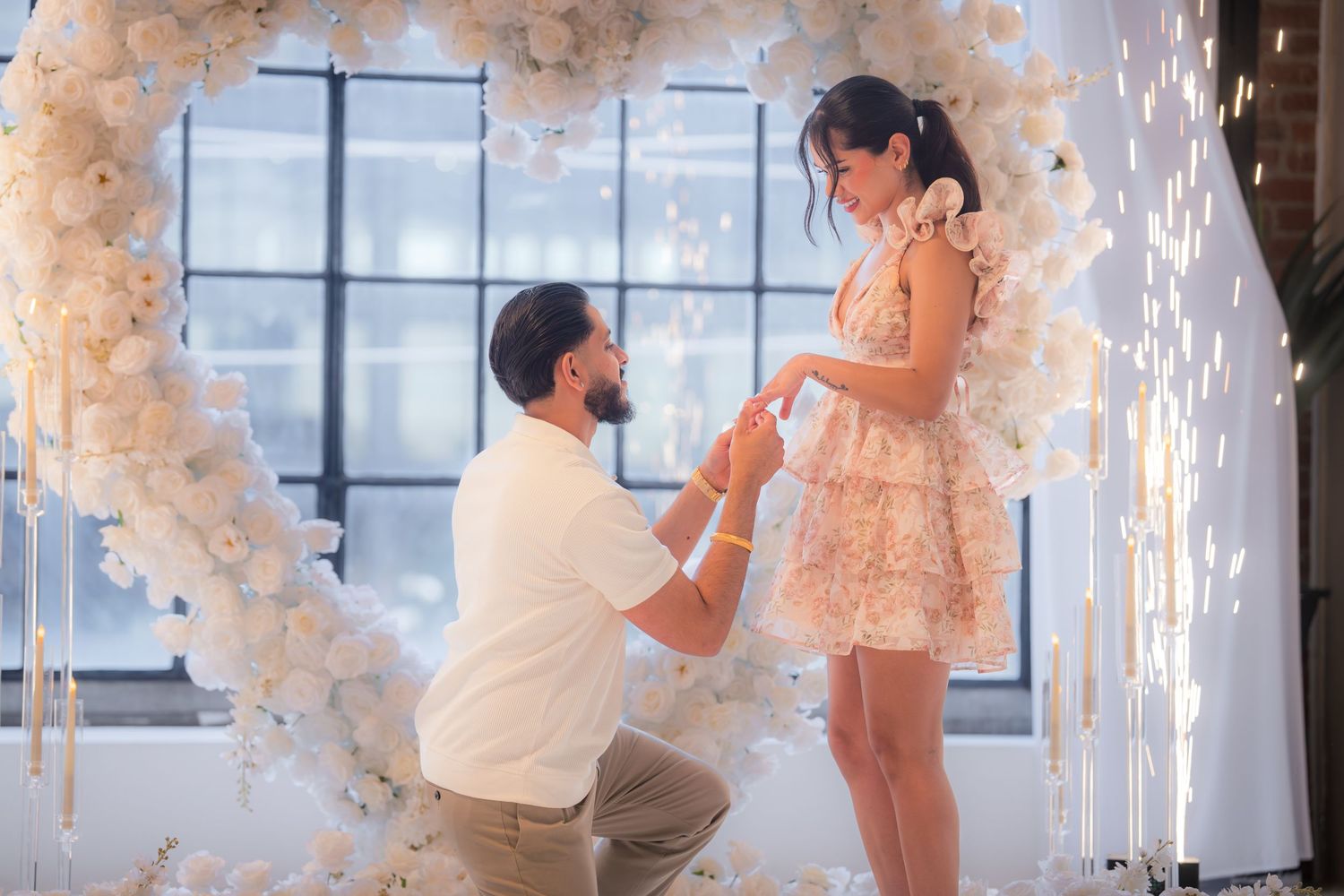 Man proposing on one knee to woman in front of white floral heart arch.