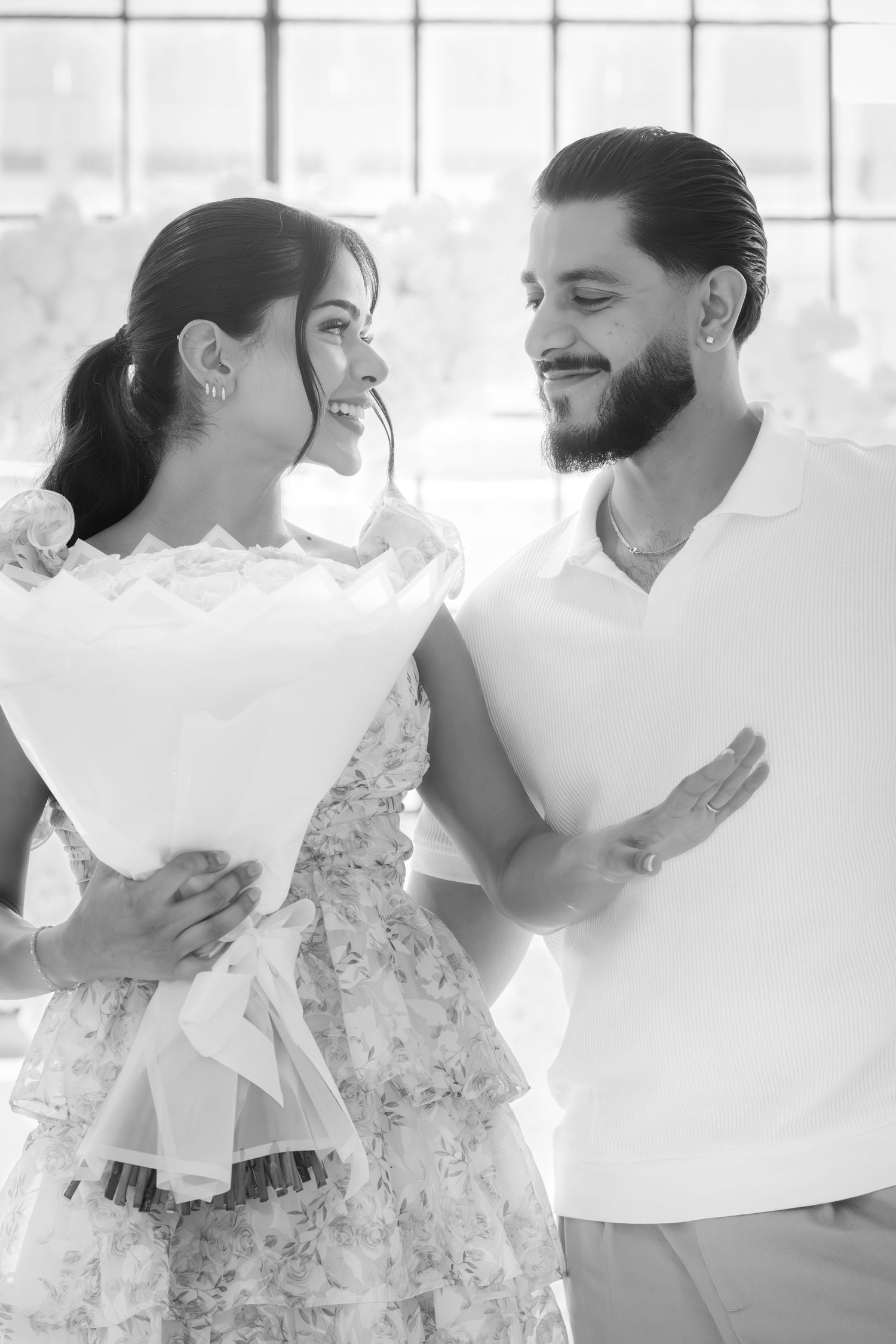 Black and white photo of a couple smiling at each other, woman holding a white bouquet.