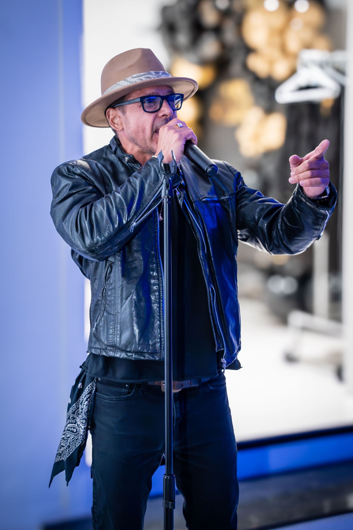 A performer in a leather jacket and hat sings into a microphone on stage.