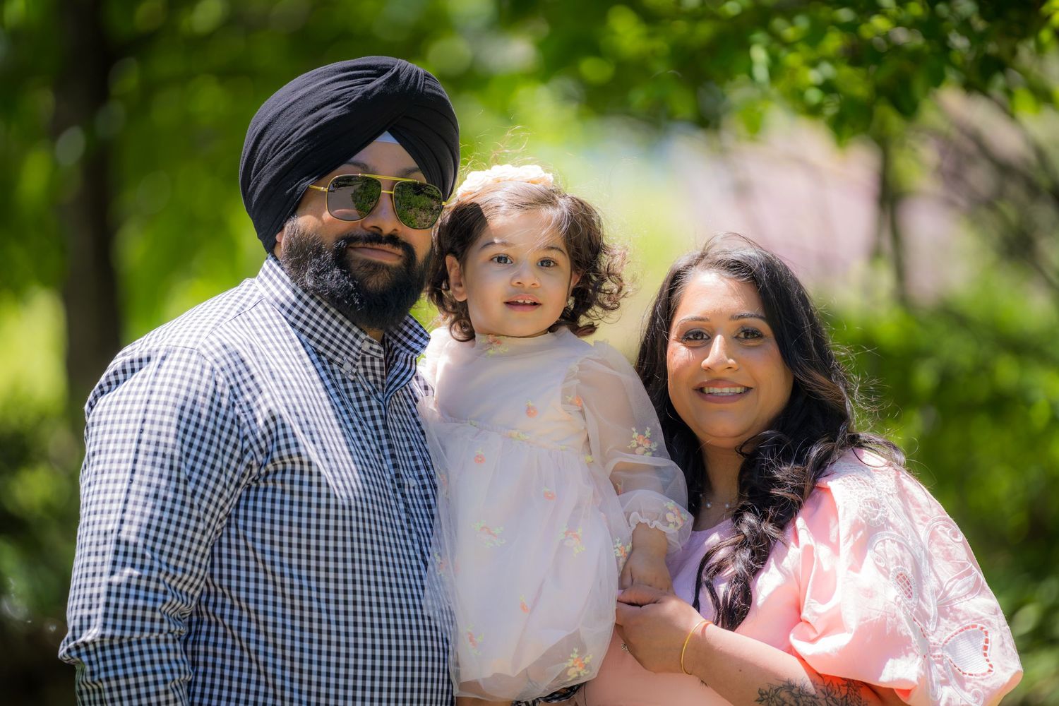 A Sikh man in turban and his wife in pink hold their young daughter in a lush green park.