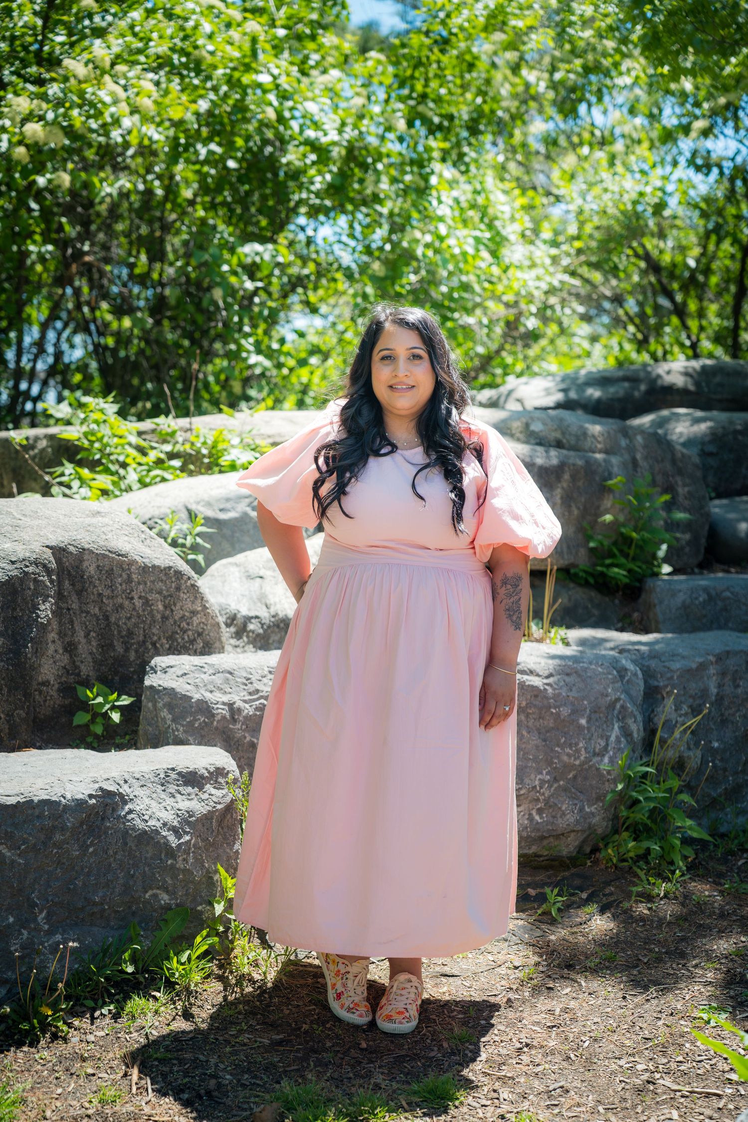 Woman in pink maxi dress with puff sleeves poses outdoors among rocks and greenery.