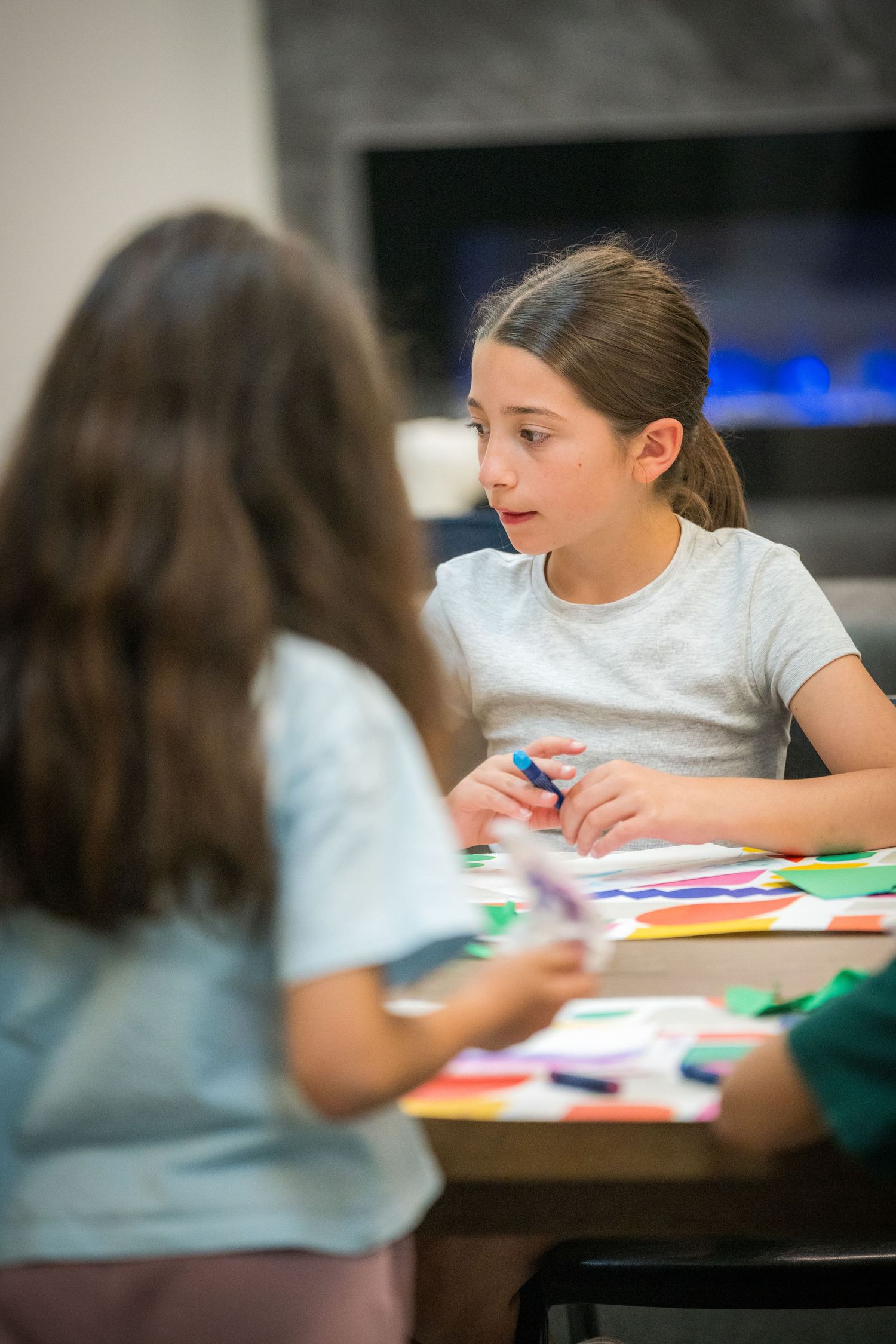 Young girl holding a marker talks with a classmate during a colorful paper craft art activity at a table.