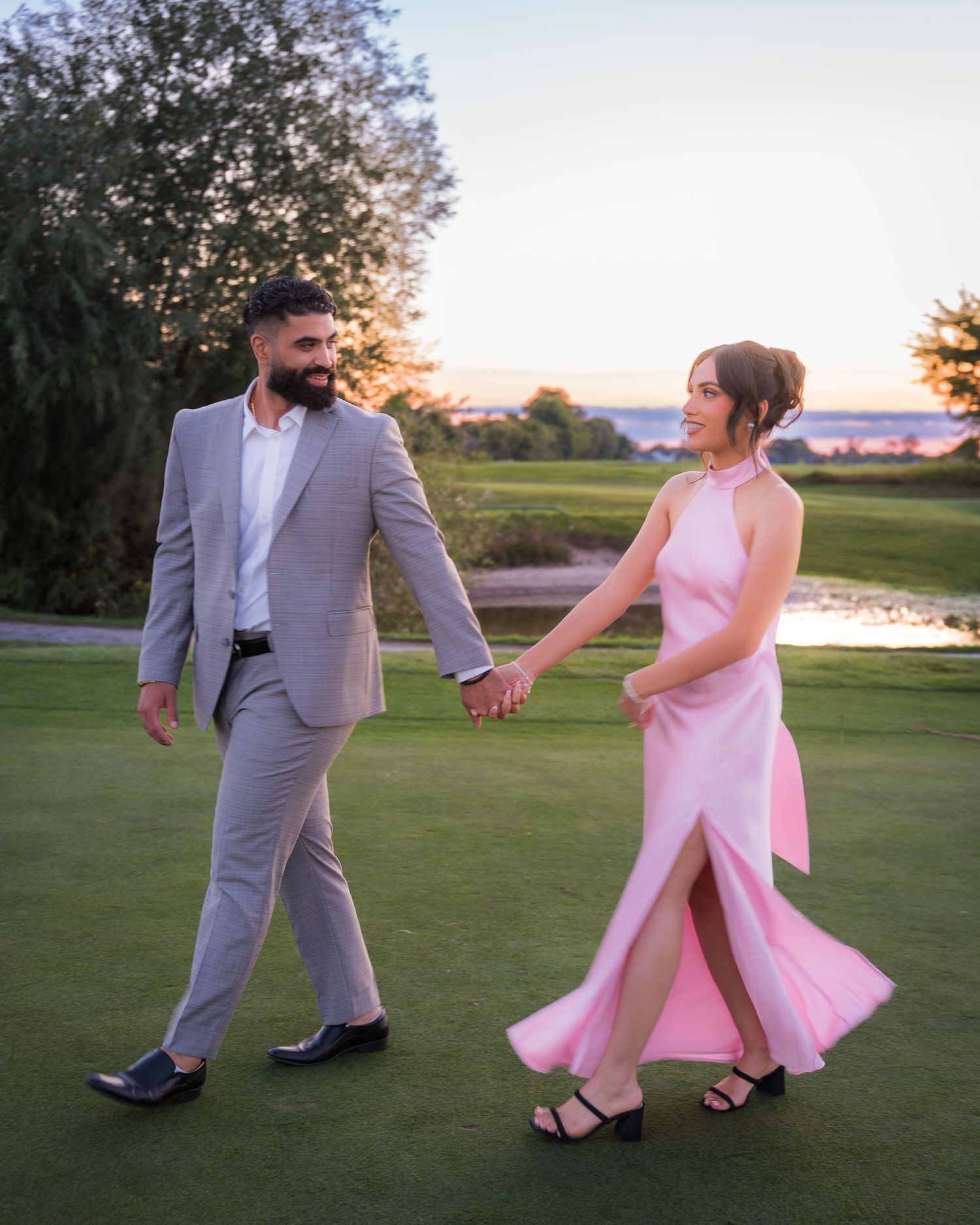 Couple in pink dress and gray suit playfully dancing on a golf course at sunset.