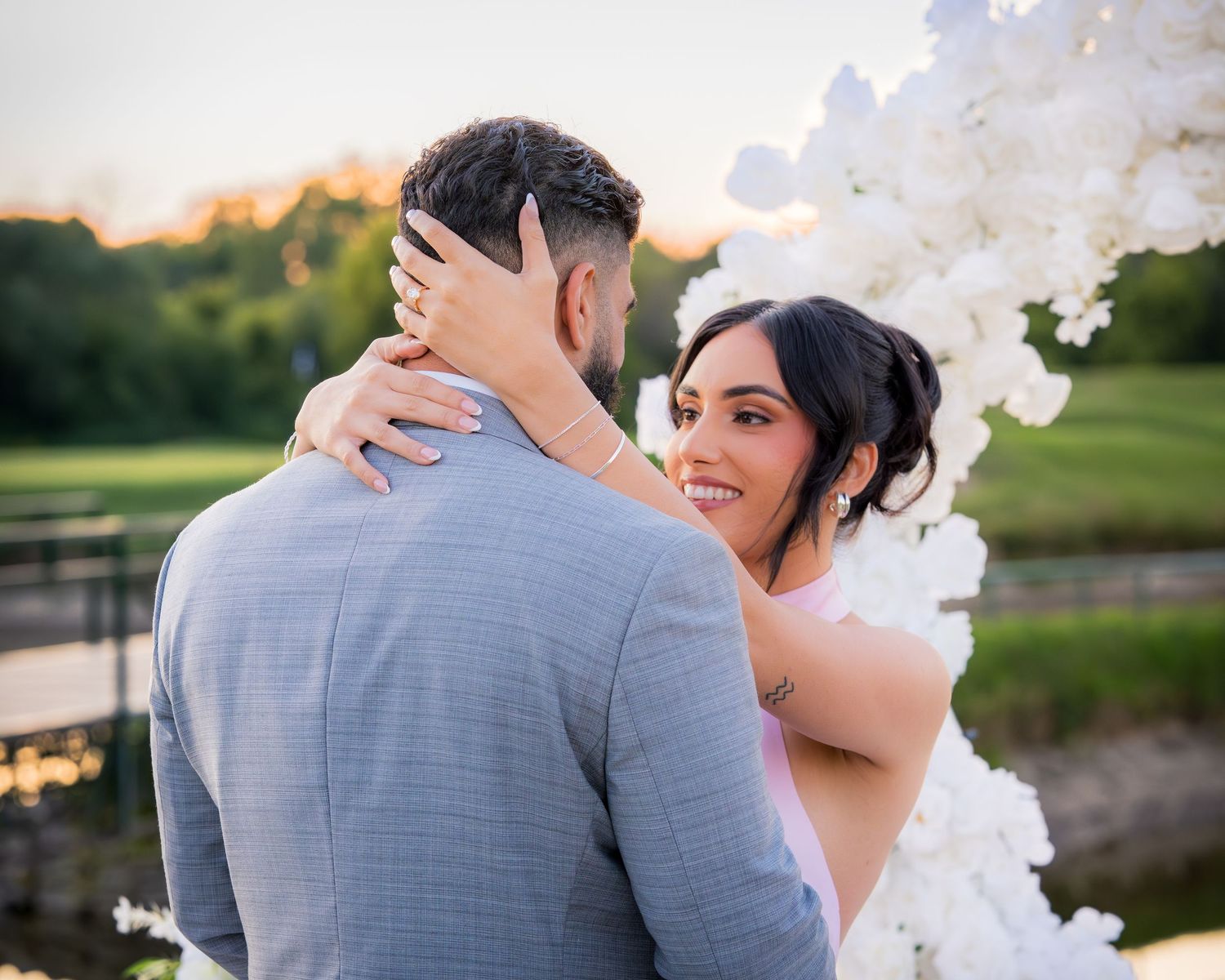 A romantic moment under a white floral arch at sunset during an outdoor wedding ceremony.