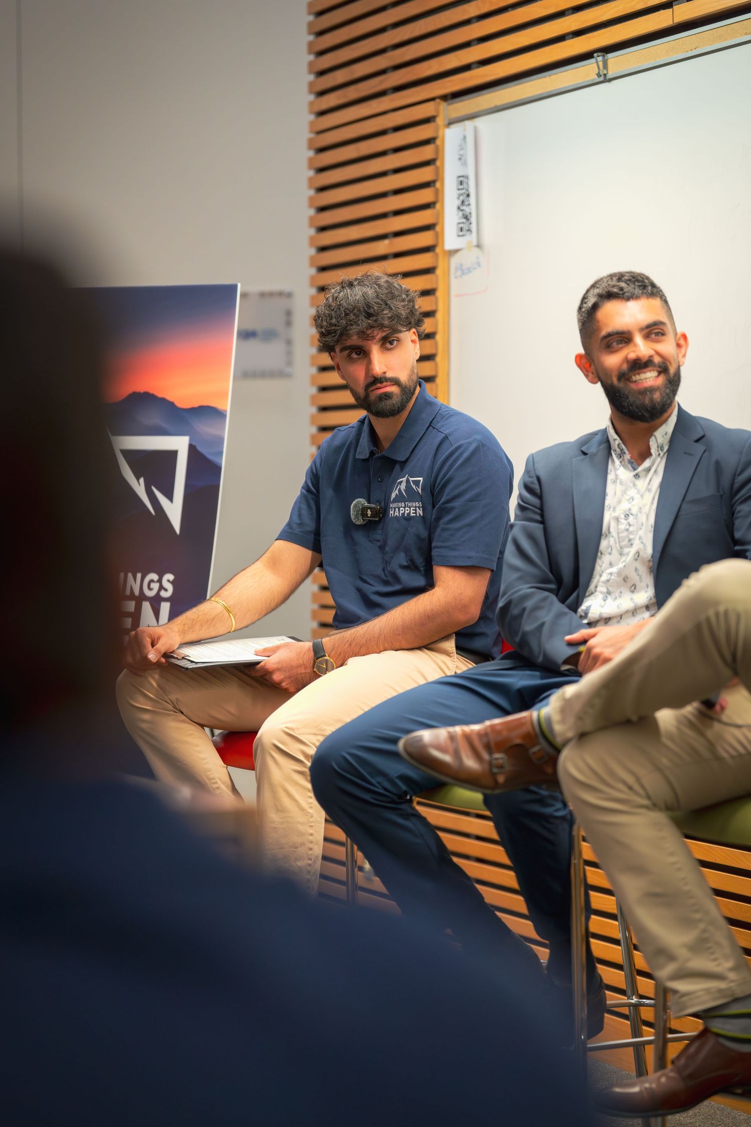 A speaker in a blue polo gestures while seated during a presentation in a meeting room.