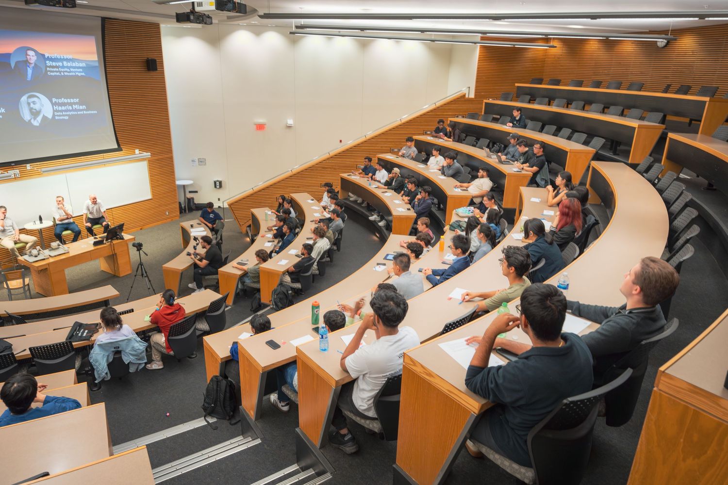 Wide angle view of a modern lecture hall with curved seating rows and presentation screen.