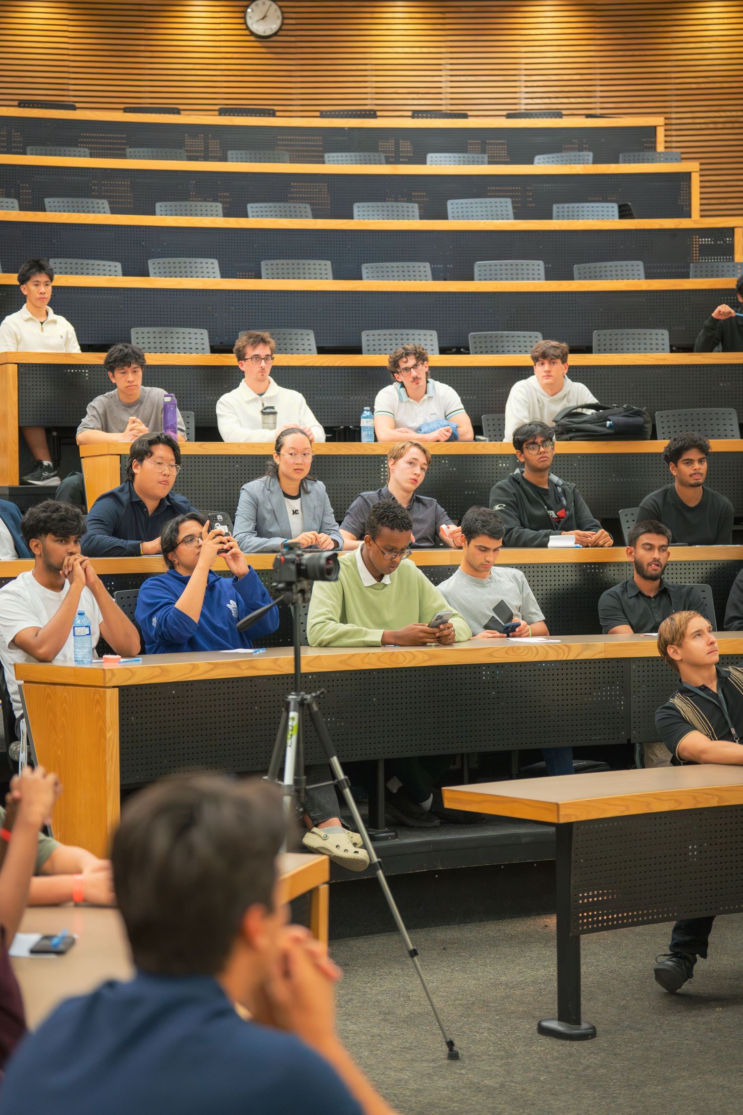 Students listening attentively in a tiered lecture hall with wooden architectural details.