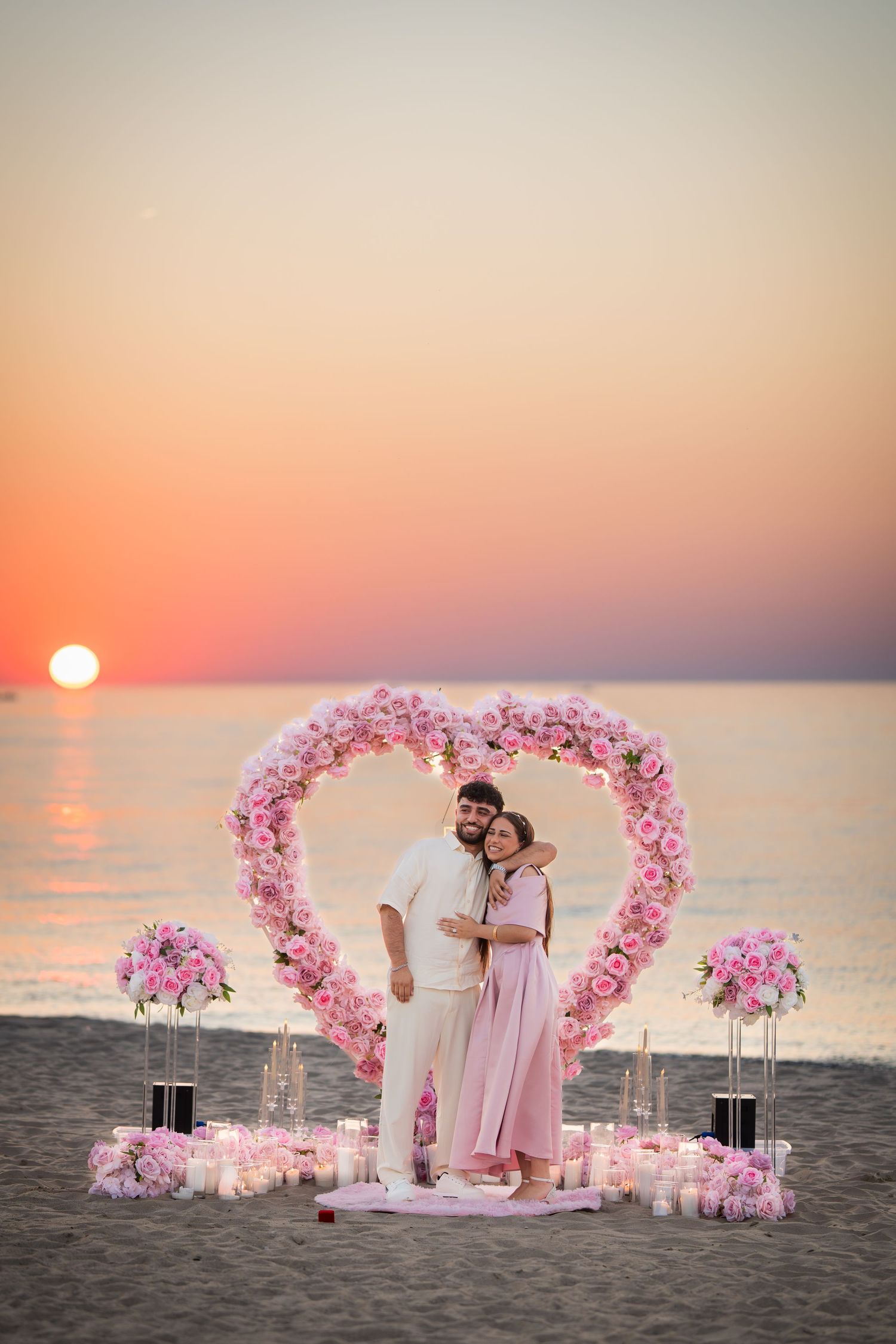 Romantic beach proposal setup with pink floral heart arch and candles against an orange sunset sky.