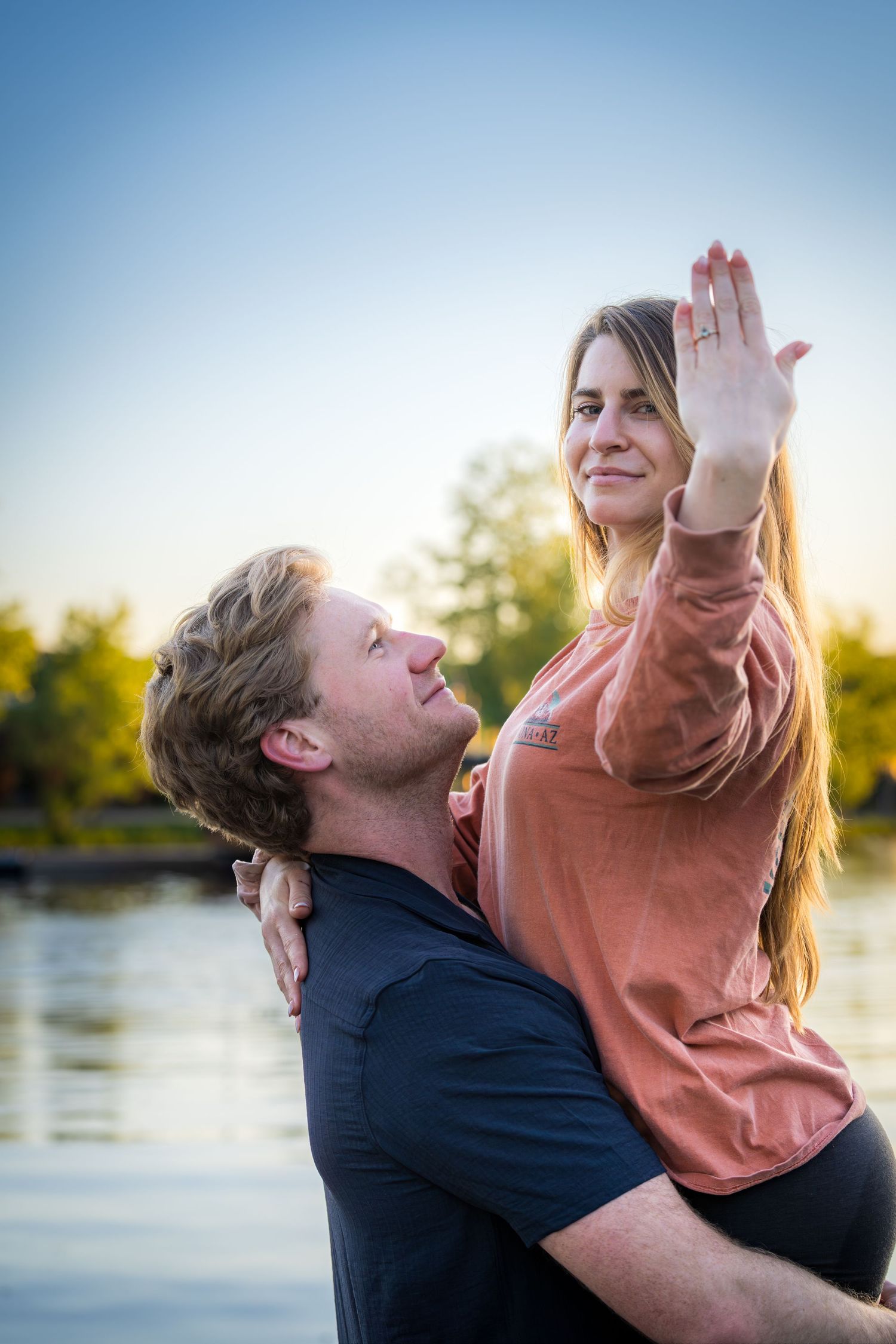 Man lifting woman by a lake at sunset while she raises her hand showing an engagement ring.